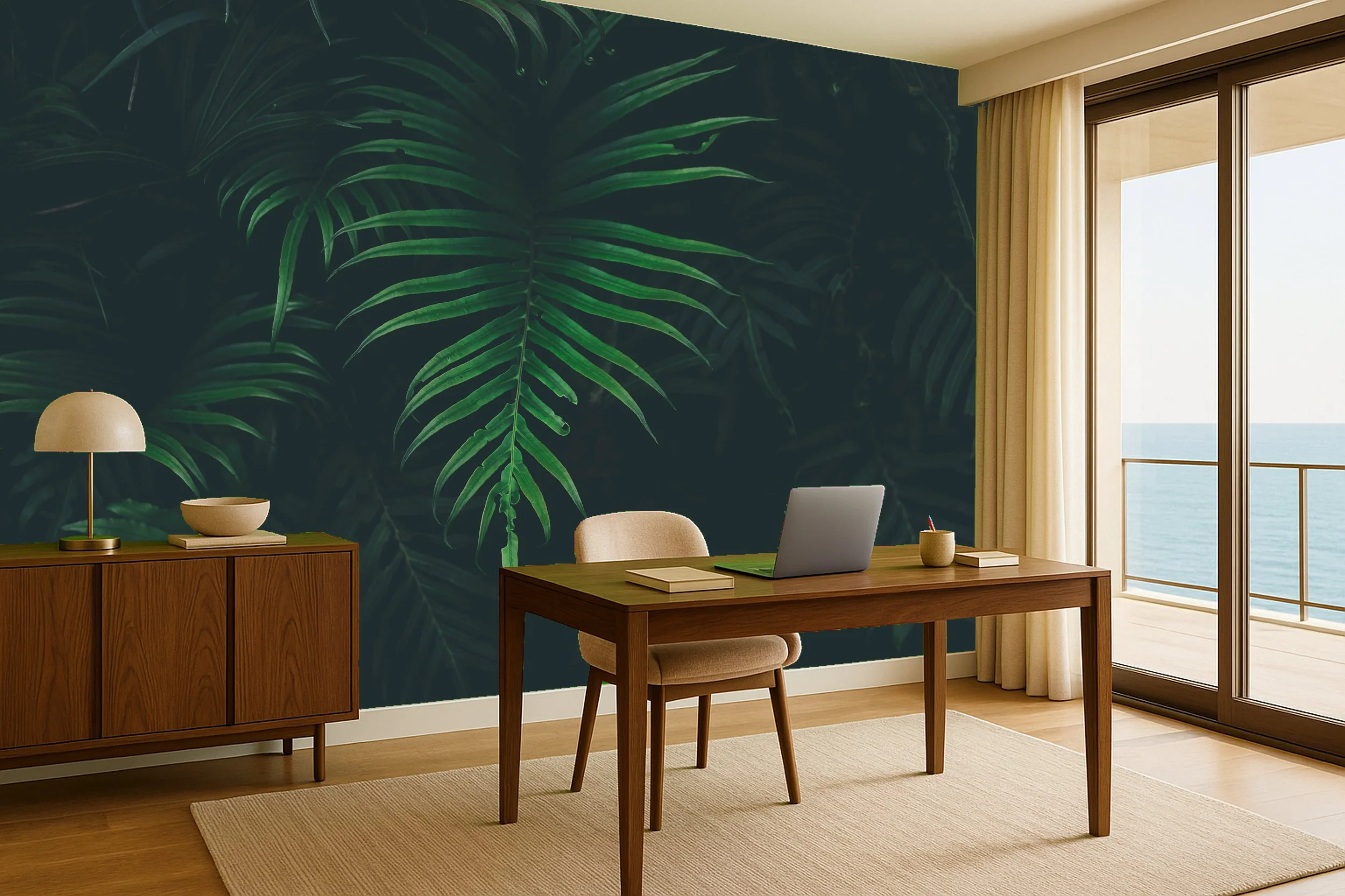 A home office with a wooden desk, beige chair, laptop, books, and a pencil holder. There is a wooden sideboard with a lamp, a bowl, and a book. A green tropical leaf mural is on the wall. Large glass door with beige curtains opens to a balcony overlooking the ocean.