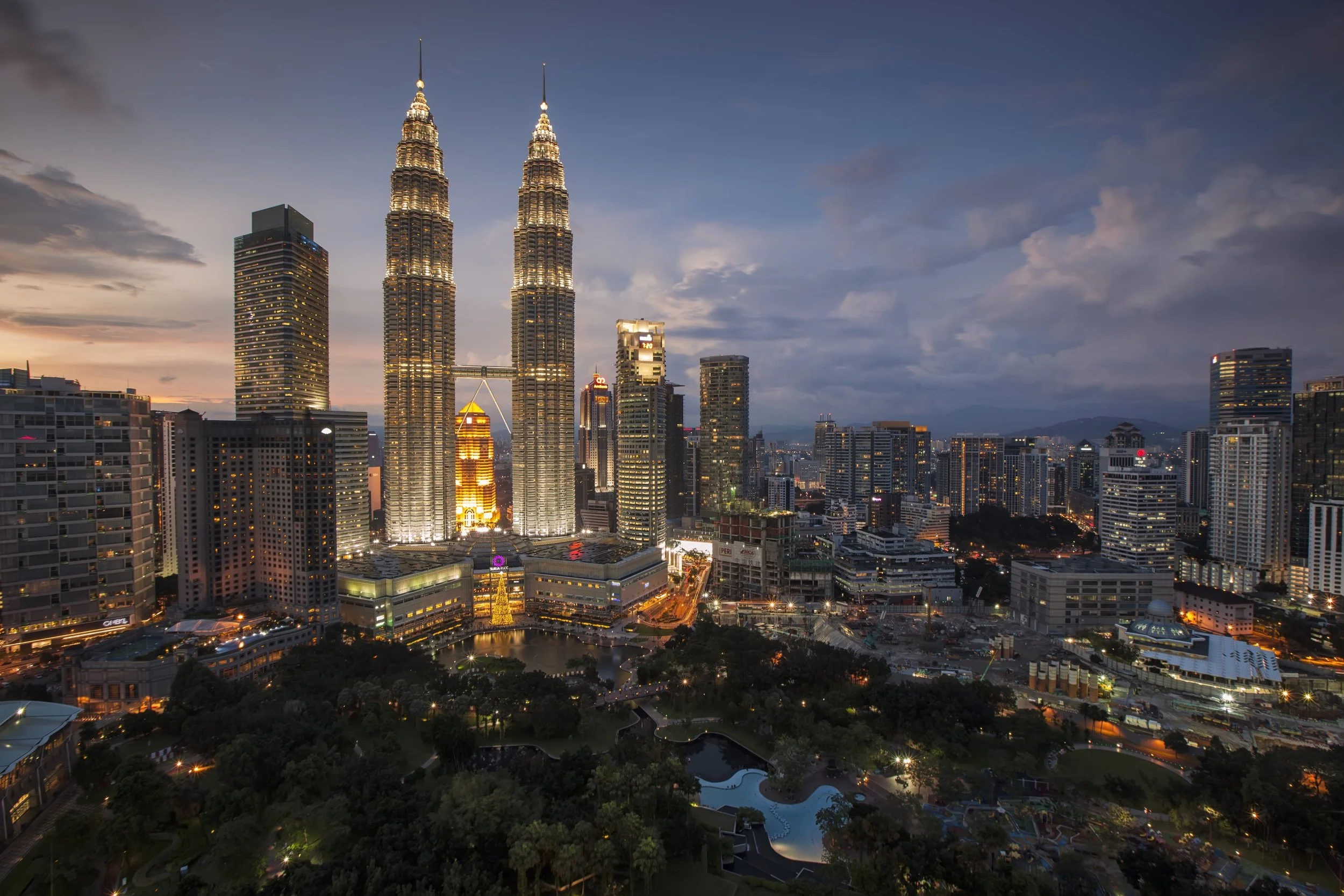 Nighttime cityscape of Kuala Lumpur featuring the illuminated Petronas Twin Towers skyline with surrounding high-rise buildings and greenery.