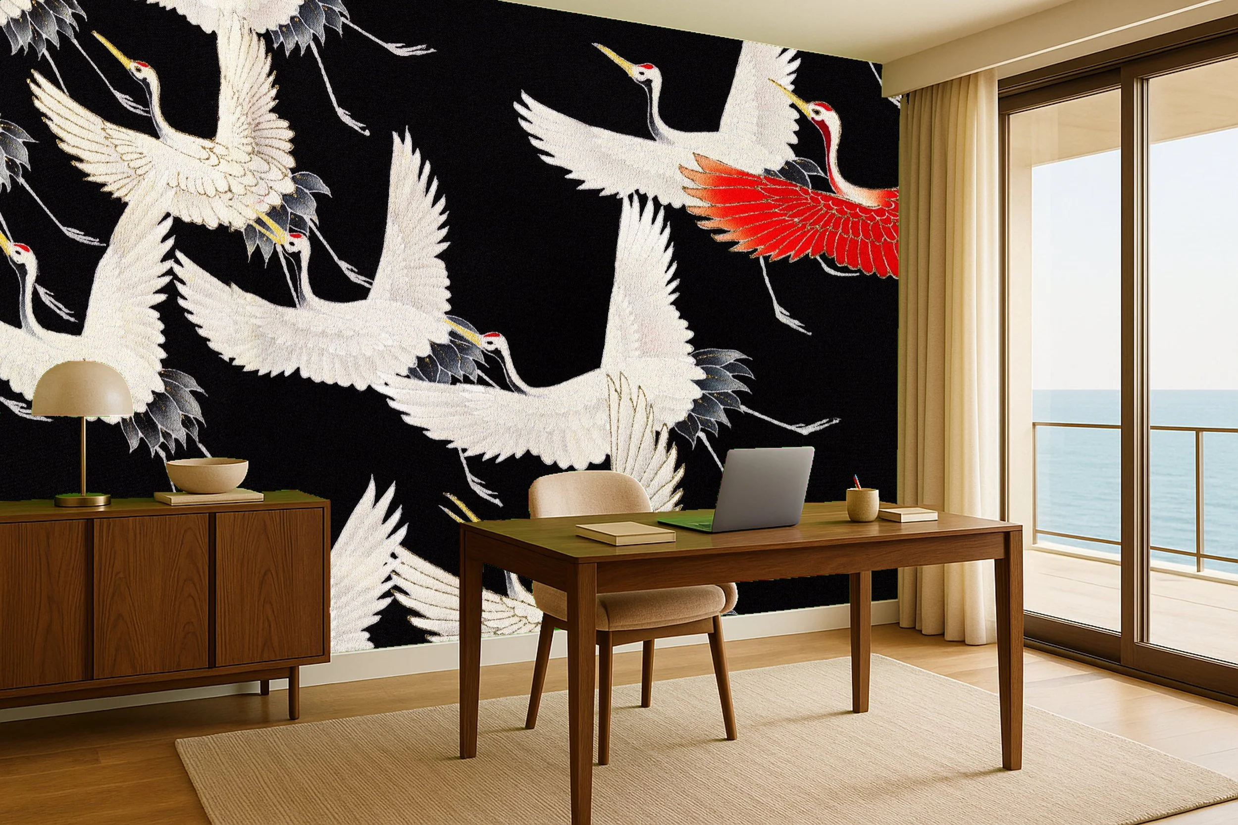 Room with a wooden desk and chair, phone, laptop, notebooks, and a bowl. A sideboard with a lamp and bowl on it. A large window or sliding door with curtains shows an ocean view. The accent wall features large artwork of white and red cranes on a black background.
