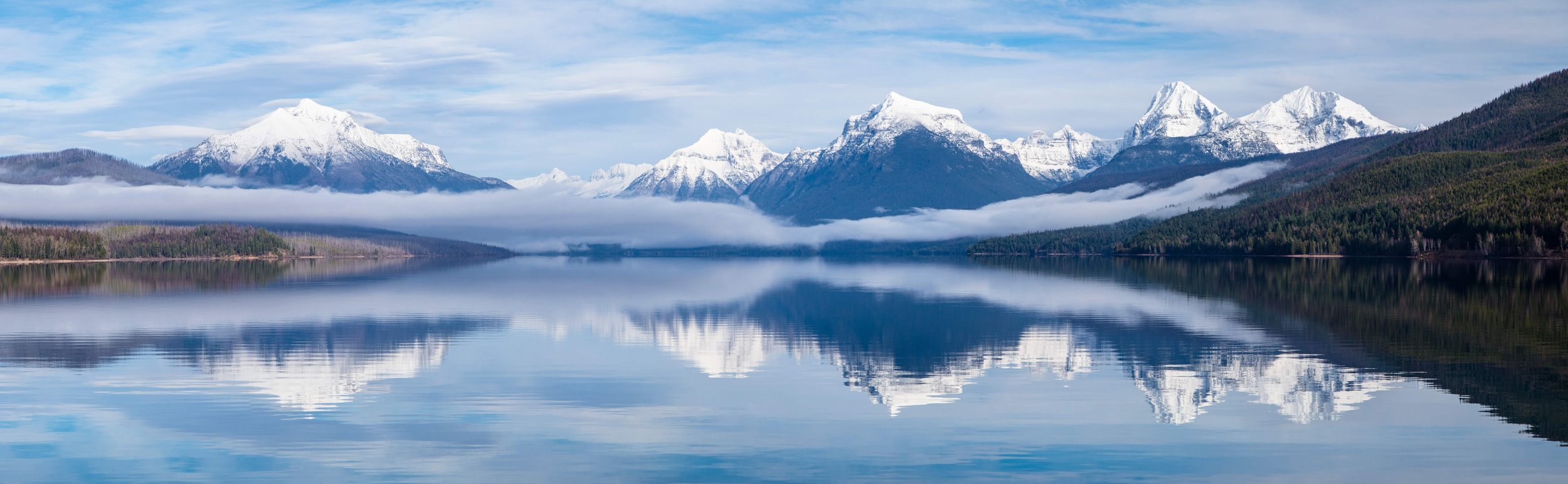 Snow-capped mountains reflected in a calm lake with green forested shoreline and a sky with scattered clouds.