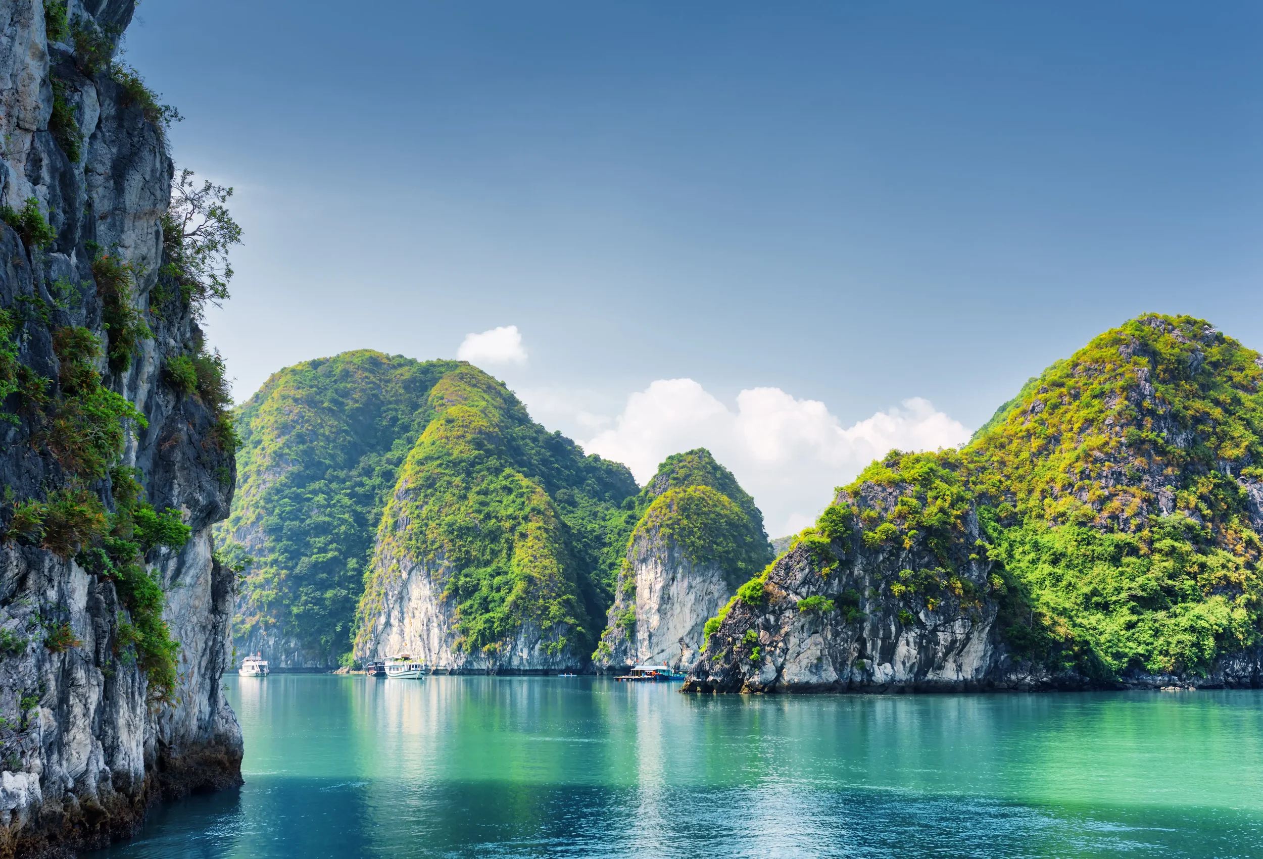 A scenic view of a bay with turquoise water, surrounded by lush green, steep limestone islands under a blue sky with white clouds.