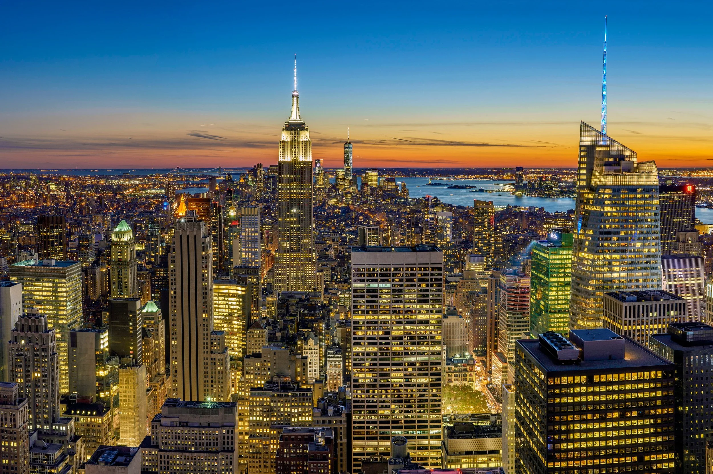 Panoramic view of New York City skyline at sunset, featuring the Empire State Building and surrounding skyscrapers, with the Hudson River in the background.