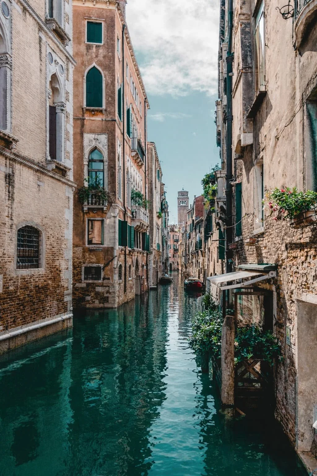 Venetian canal with historic buildings along the waterside, featuring colorful facades, green shutters, and small balconies with plants, with a clock tower visible in the distance under a partly cloudy sky.
