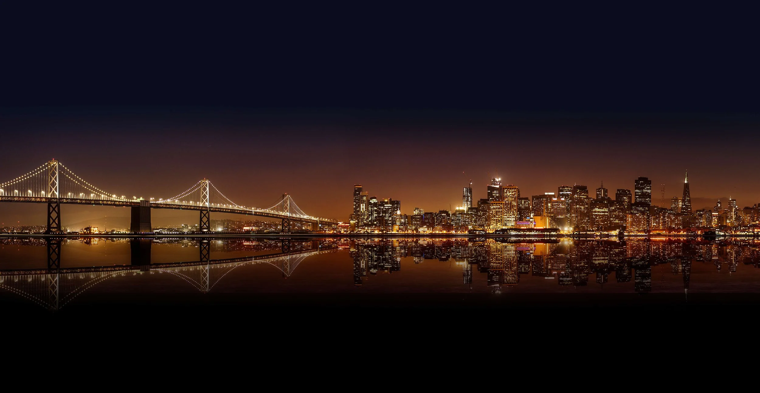 Nighttime city skyline of San Francisco with the Bay Bridge illuminated, reflected in the water.
