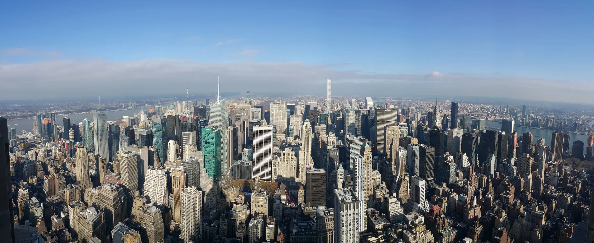 A panoramic view of Midtown Manhattan skyscrapers, including the Empire State Building, with the Hudson River and New Jersey in the background on a clear day.