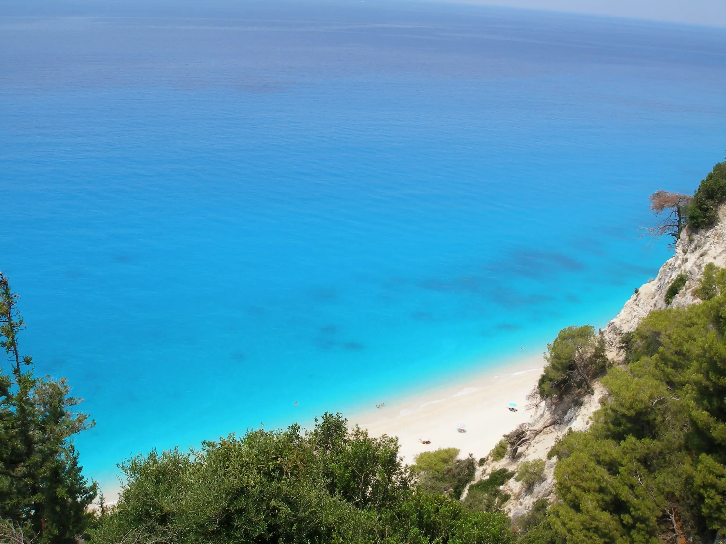 Aerial view of a turquoise blue ocean meeting a sandy beach with a few umbrellas and trees on the shoreline, surrounded by green vegetation and white cliffs.