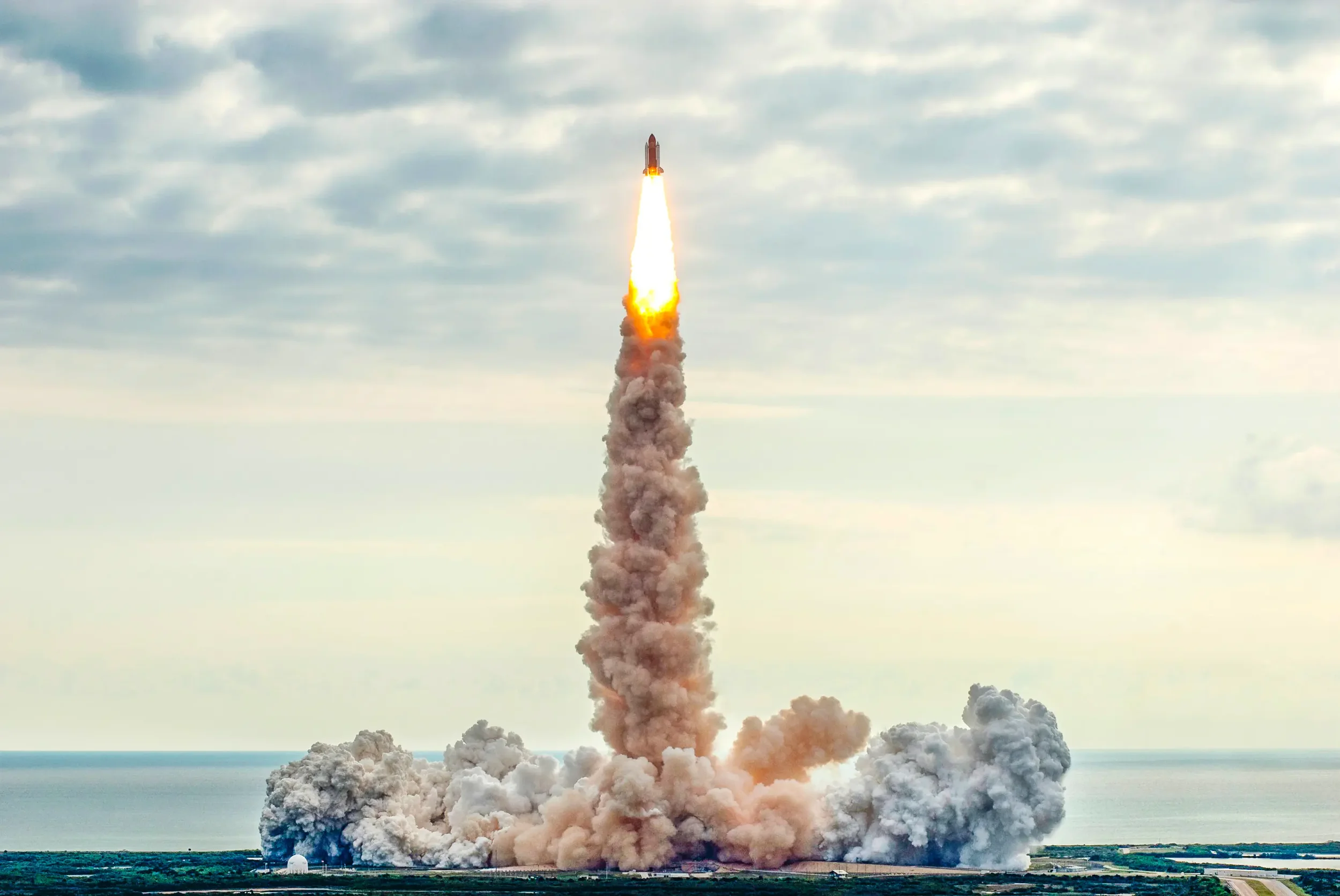 A rocket launches into the sky, leaving a large plume of smoke and fire behind it, with an ocean and cloudy sky in the background.