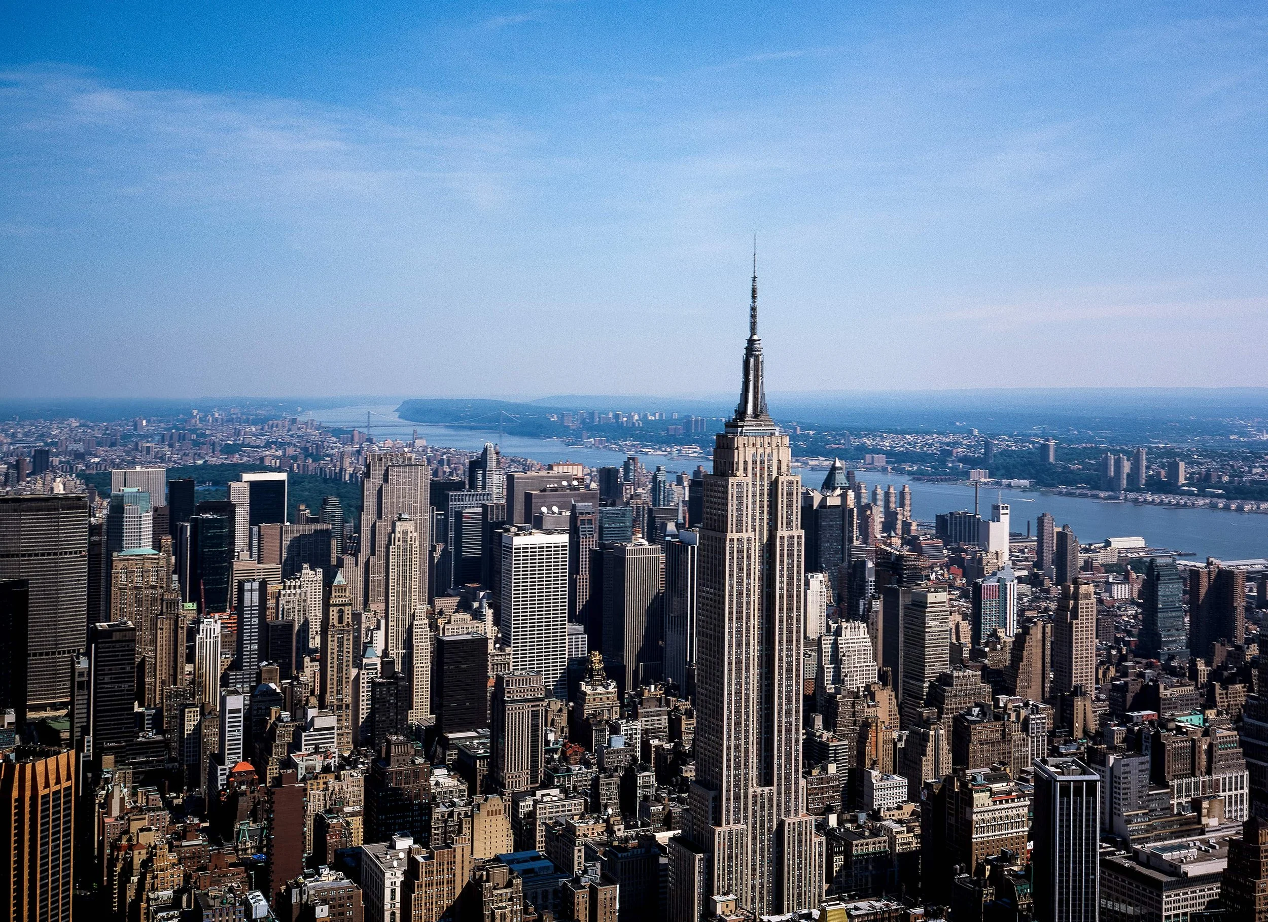 Aerial view of New York City skyline featuring the Empire State Building, with other skyscrapers and a river in the background on a clear day.