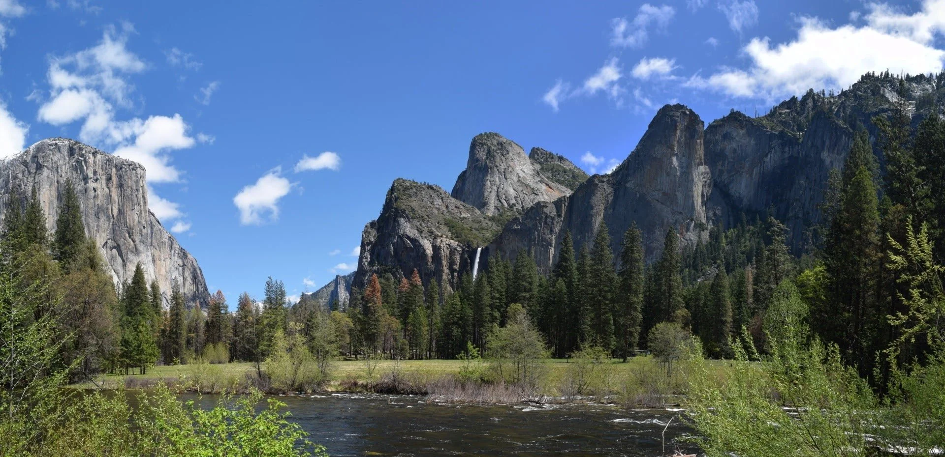 Scenic view of rocky mountains with green forests and a river flowing in the foreground under a partly cloudy blue sky.
