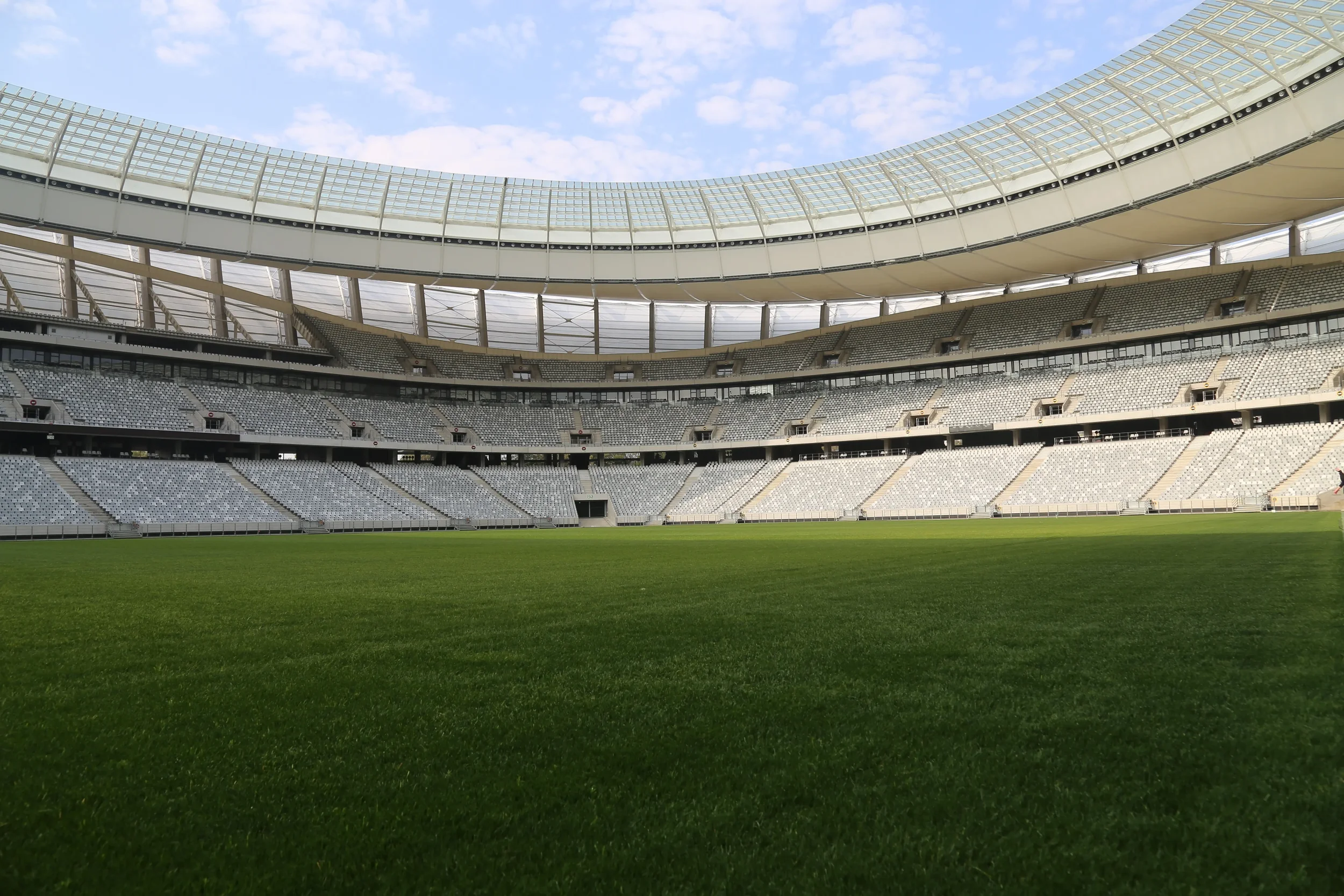Empty stadium with green grass field, surrounded by multiple tiers of seats under a bright sky.