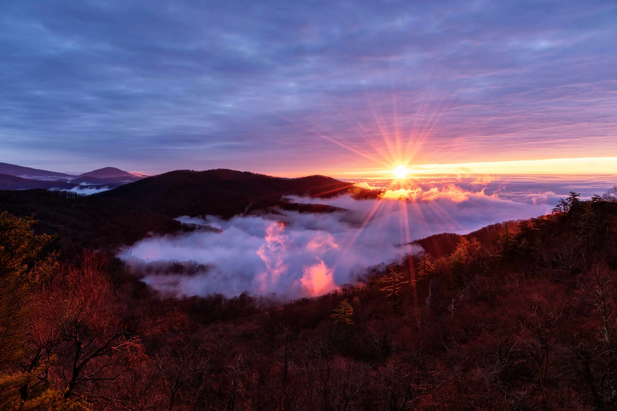 Sunrise over misty mountains with colorful sky, clouds, and trees in the foreground.