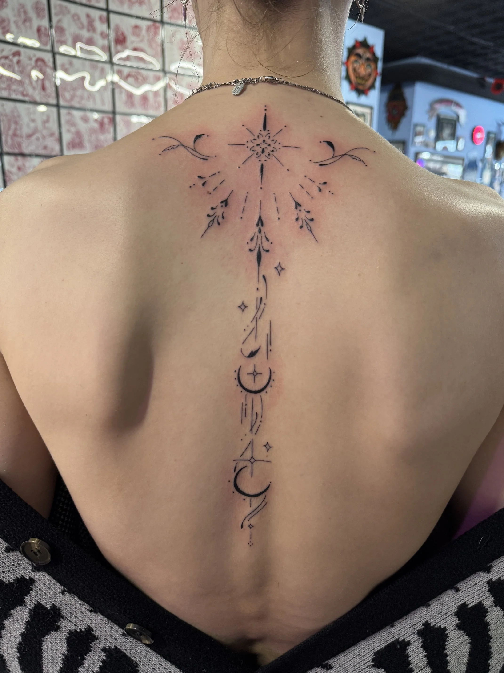 Tattoo on upper back and neck, featuring a central starburst or compass design, with crescent moons, stars, and delicate linework extending down the spine.
