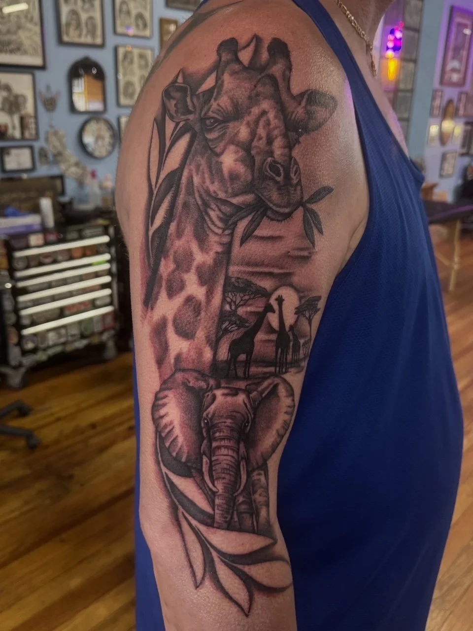 Tattoo of a giraffe and an elephant on a person's arm, with a savannah landscape and trees in the background.
