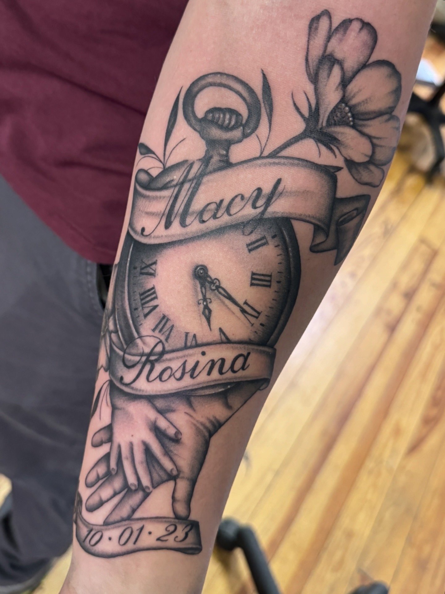 Tattoo of a clock with a banner displaying the name 'Macy', surrounded by flowers, a pair of hands, and a date '10-01-23' on the forearm.