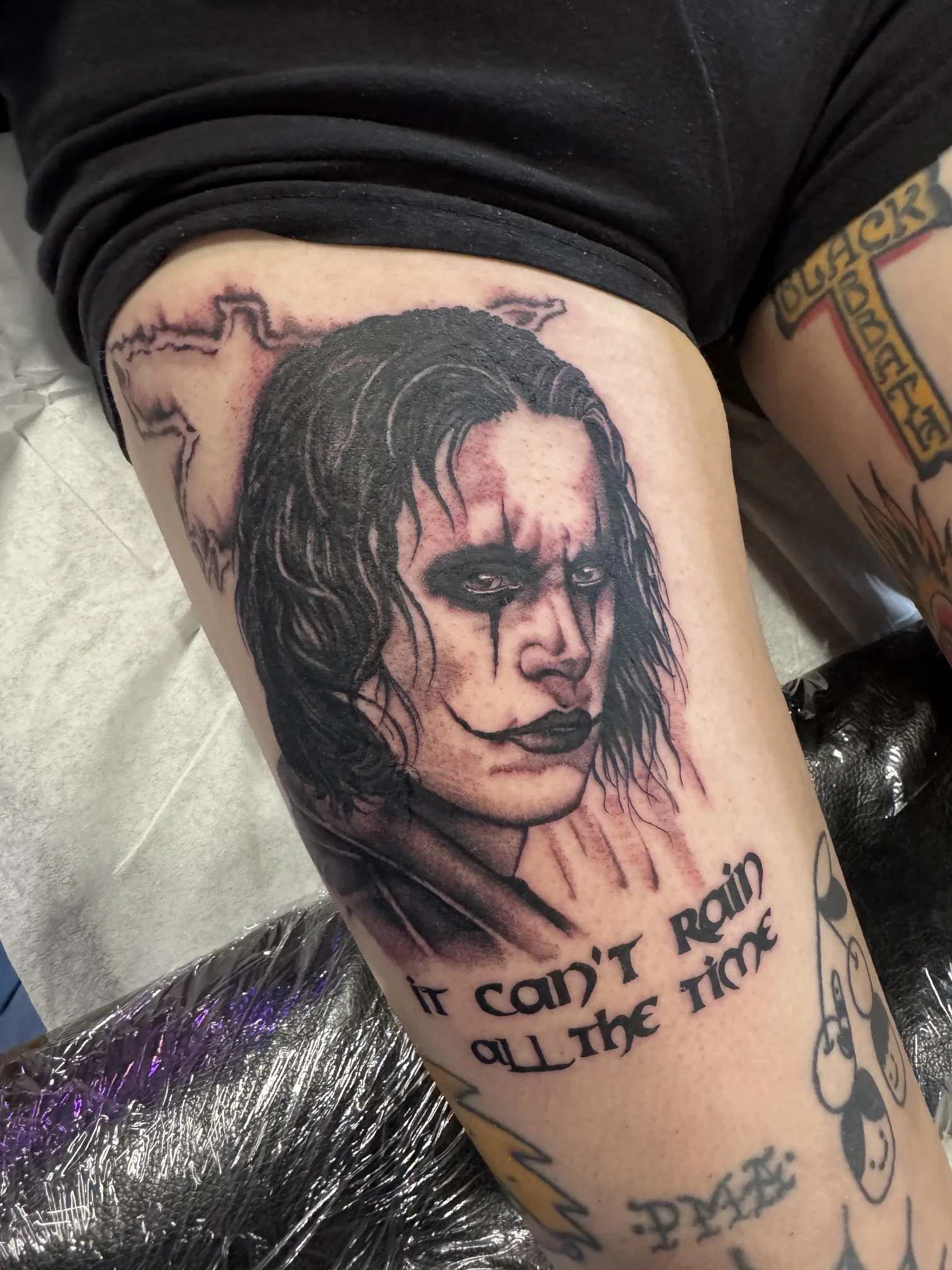 Close-up of a tattooed arm with a detailed black and gray portrait of Eric Draven, from the movie The Crow, surrounded by some text.