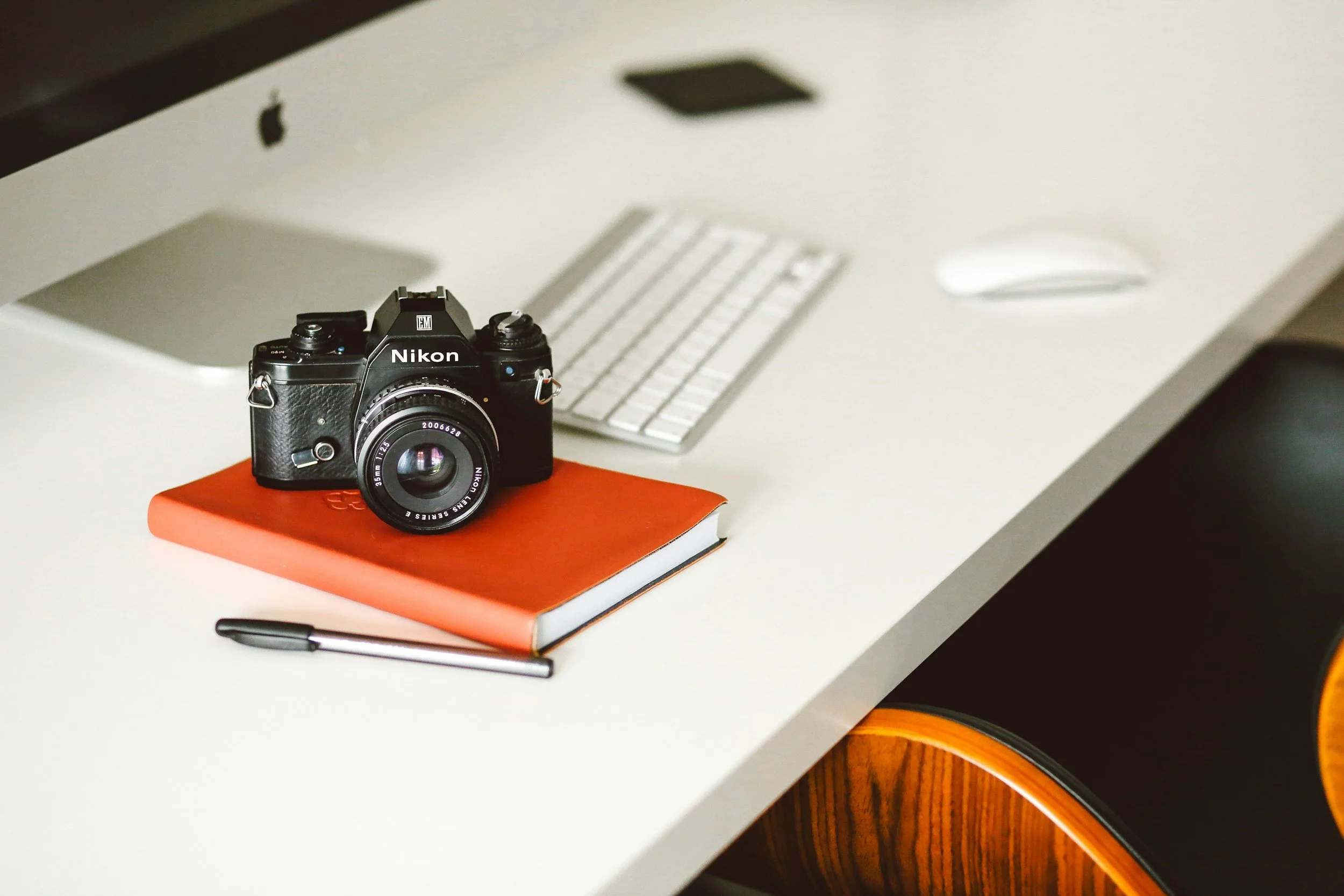 Et svart Nikon-kamera liggende på en oransje notatbok med en svart penn, på et hvitt skrivebord. I bakgrunnen står en sølvfarget Apple iMac med trådløst tastatur og mus.