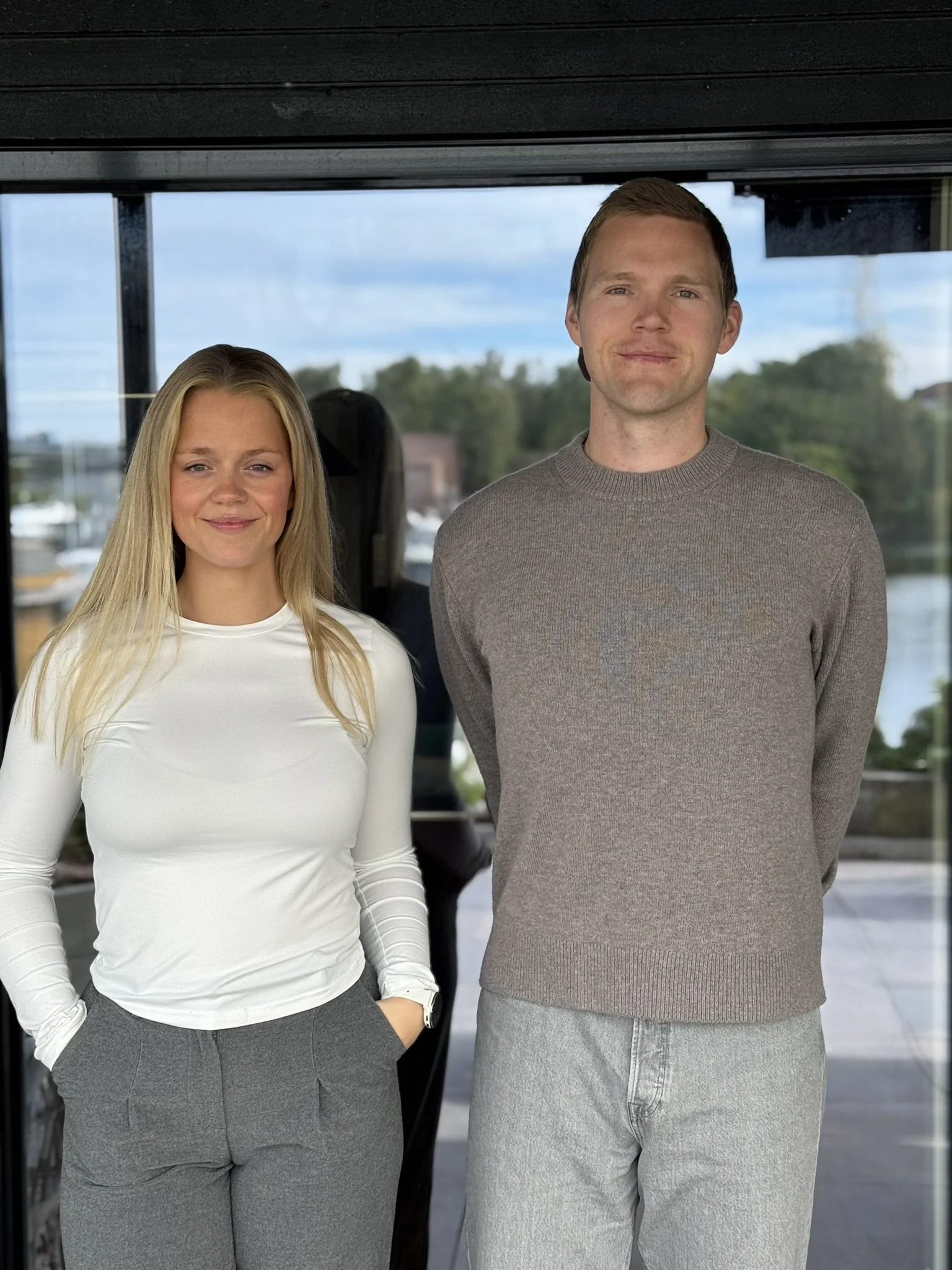 En kvinne med langt blondt hår i hvit genser og grå bukse står ved siden av en mann med kort rødt hår i grå genser og lysegrå bukse. Begge står innendørs foran glassdører med utsikt til trær og vann.