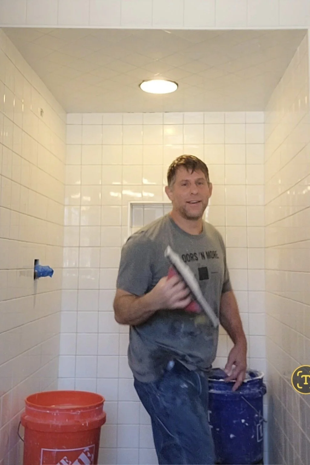 Jesse Robinson wearing a Floors ’N More shirt and holding a tile-setting tool inside a white tile shower.