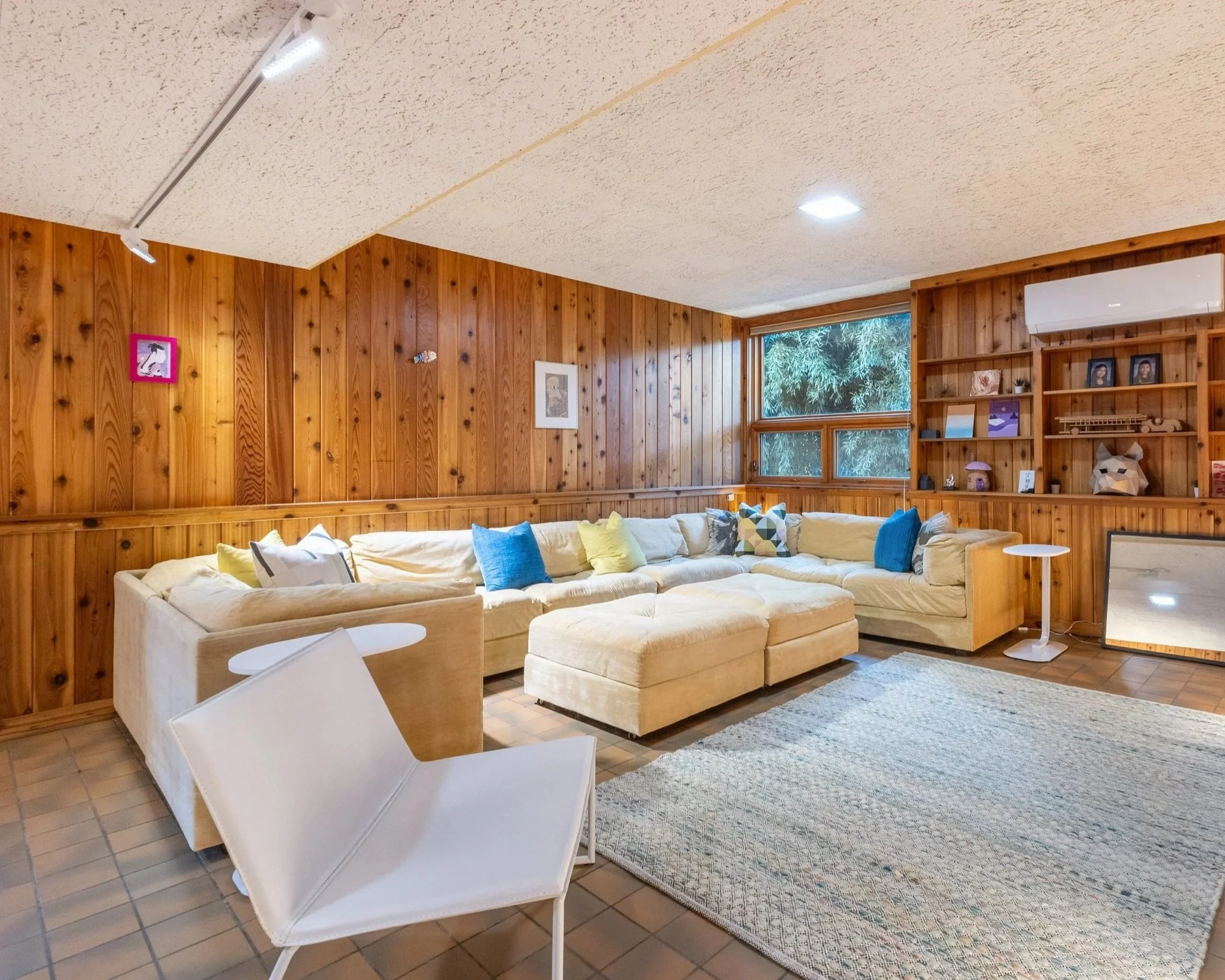 Mid-century modern lower-level family room film location with wood-paneled walls, tiled floor, large windows with natural light, and open lounge layout.