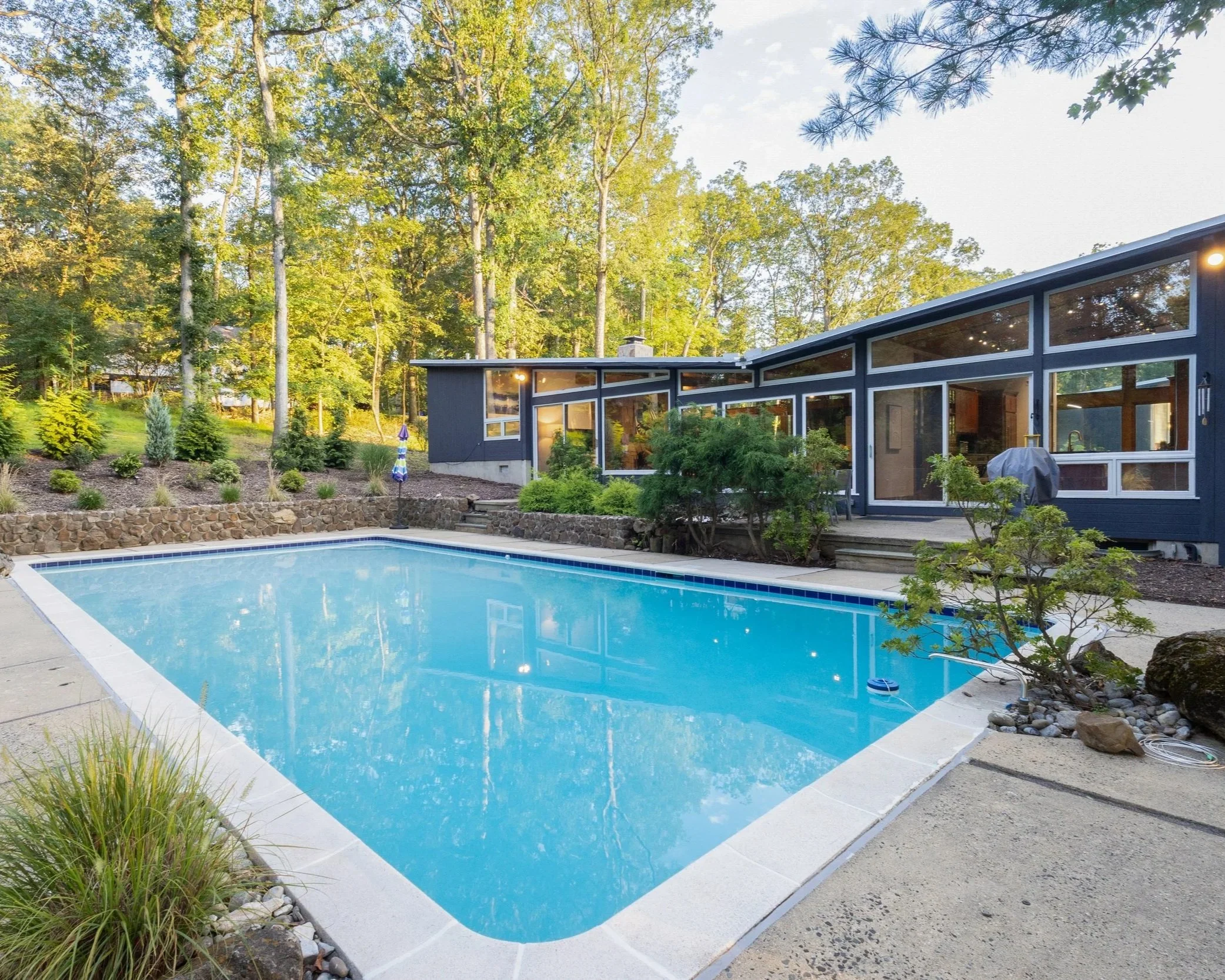 Mid-century modern filming location in New Jersey with large backyard pool, naturalistic landscaping, deck access, and glass walls reflecting wooded views.