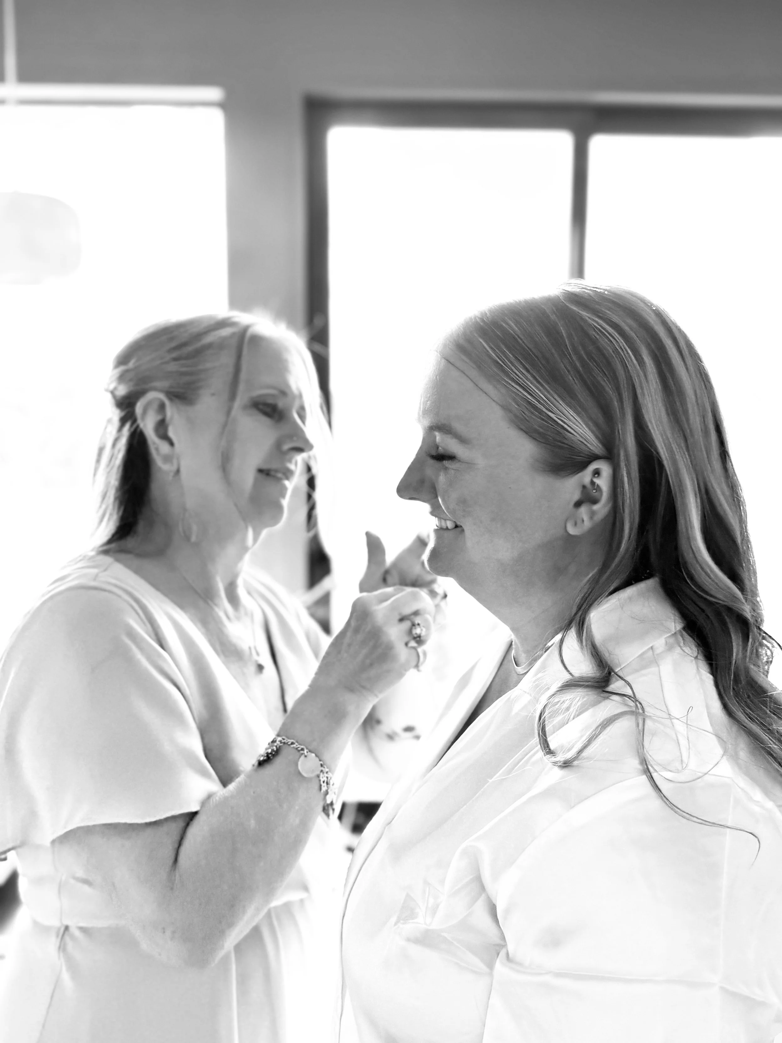 A woman with long hair getting makeup applied by a makeup artist, both smiling and facing each other, in a bright room.