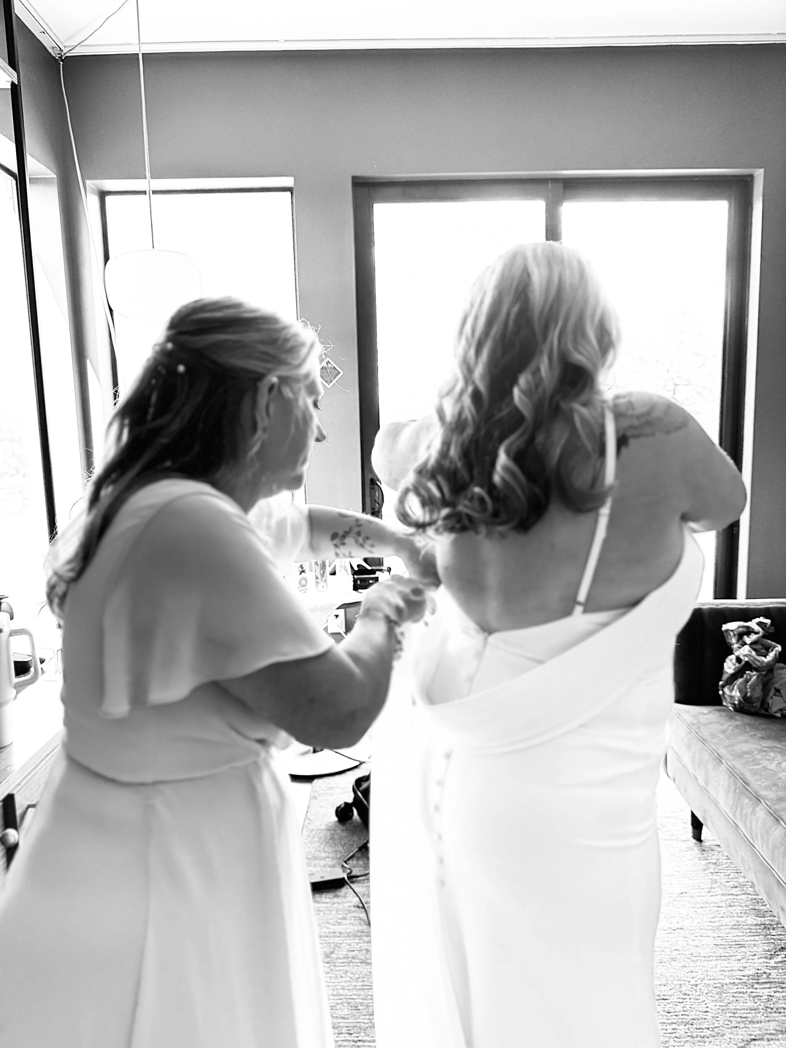 A woman helping another woman into a wedding dress, with both wearing white dresses, in a well-lit room near a window.