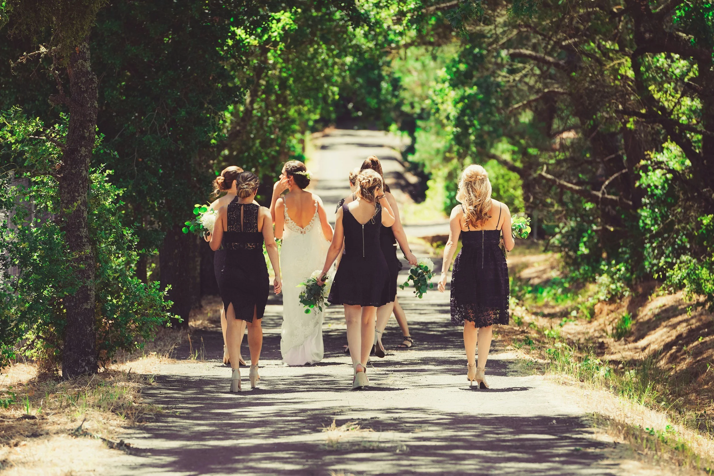 A group of women, some in black dresses and one in a white wedding dress, walking on a forested path carrying bouquets of flowers.