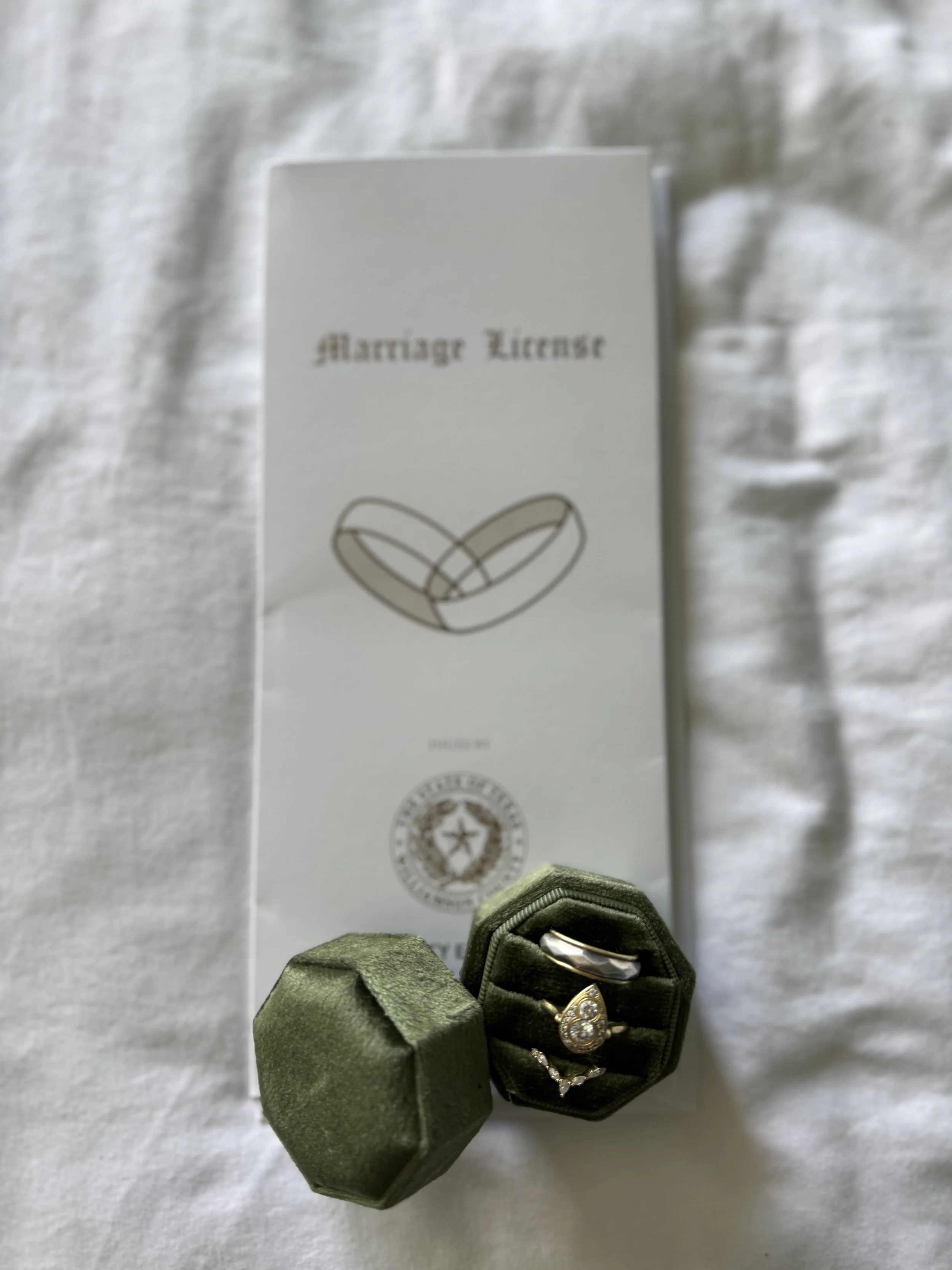A marriage license document with a simple line drawing of interlocked wedding rings on it, accompanied by a green velvet ring box containing a pearl and diamond ring, placed on a white fabric background.