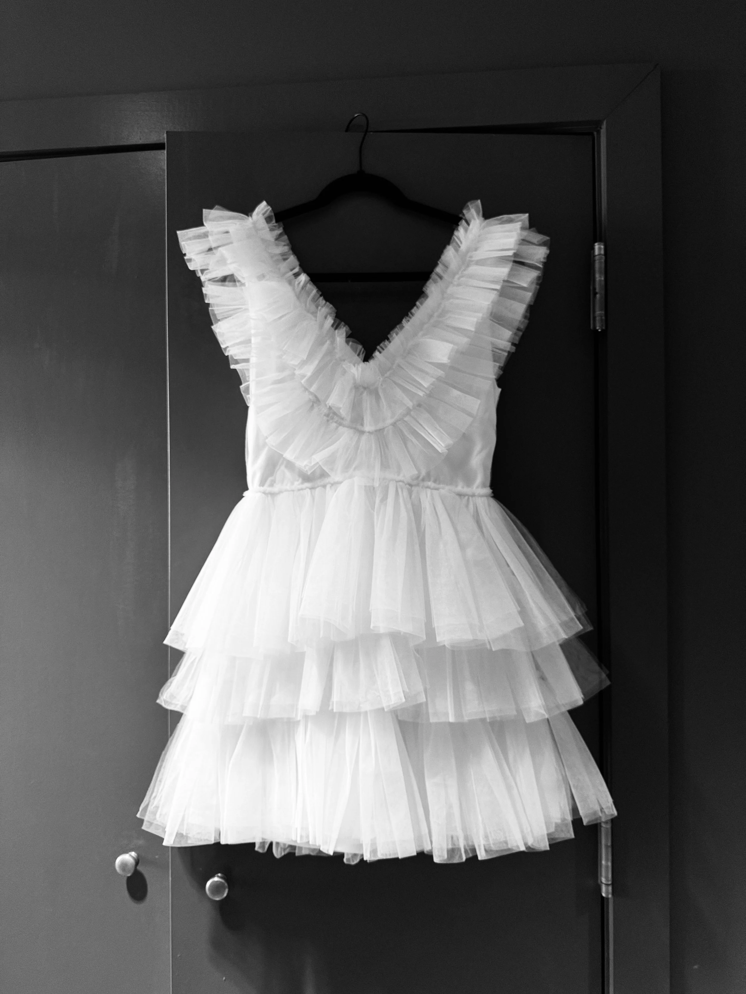 A white dress with layered ruffles hanging on a black hanger against a dark door.