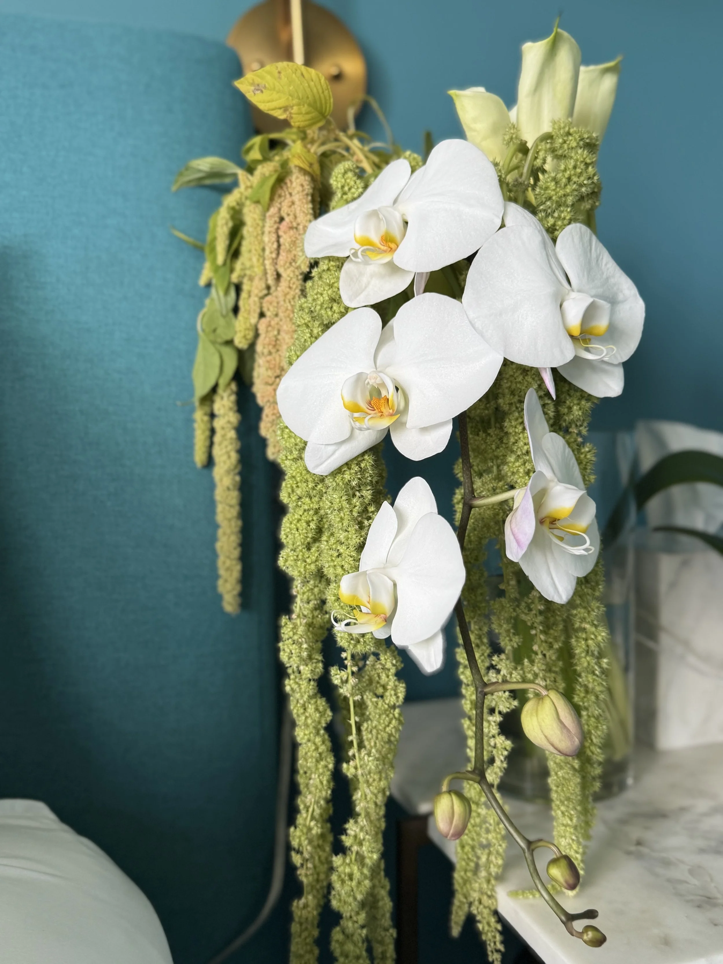 A floral arrangement featuring white orchids with yellow and pink accents, hanging green amaranthus, and light green flower buds in a glass vase on a marble surface.