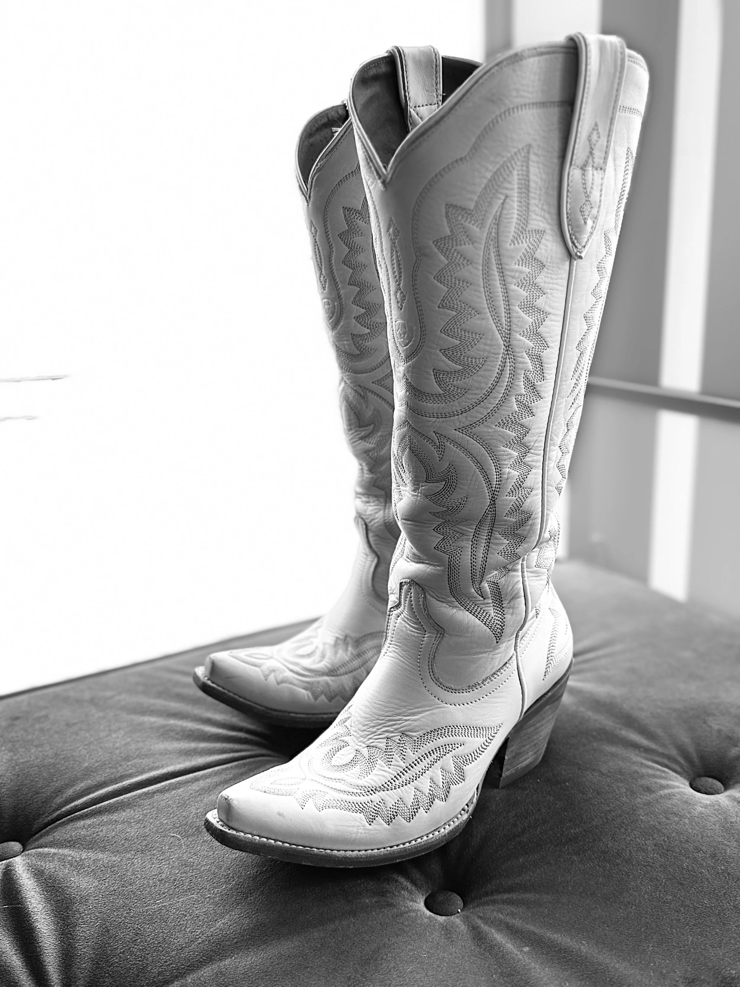 A pair of white cowboy boots with decorative stitching, placed on a tufted black leather surface.