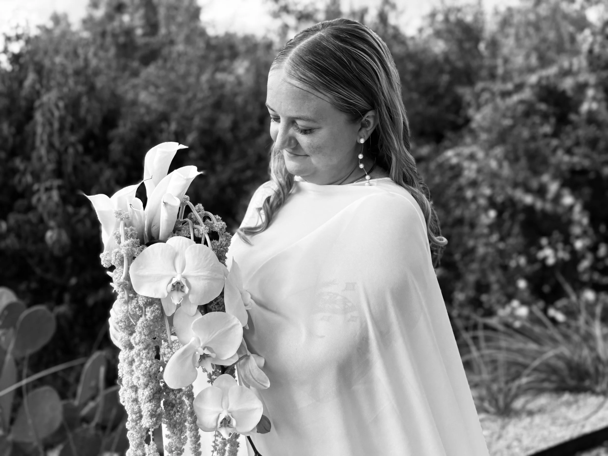 A woman with wavy hair and earrings holding a large bouquet of flowers, including orchids and calla lilies, outdoors in a garden or park setting.