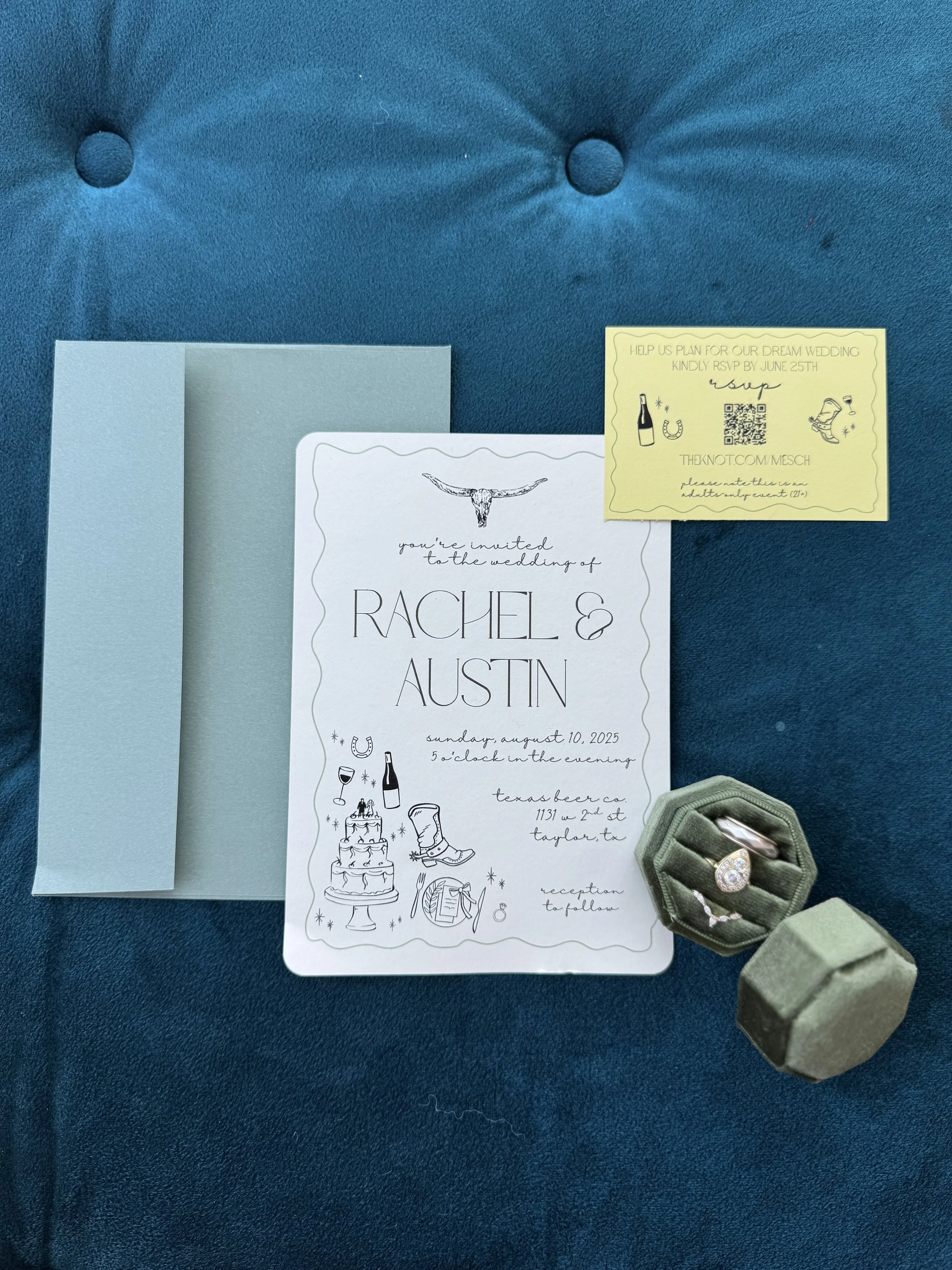 Wedding invitation for Rachel and Austin, featuring a longhorn skull illustration, with a blue envelope, a yellow RSVP card, and a green ring box with rings, all on a blue cushioned surface.