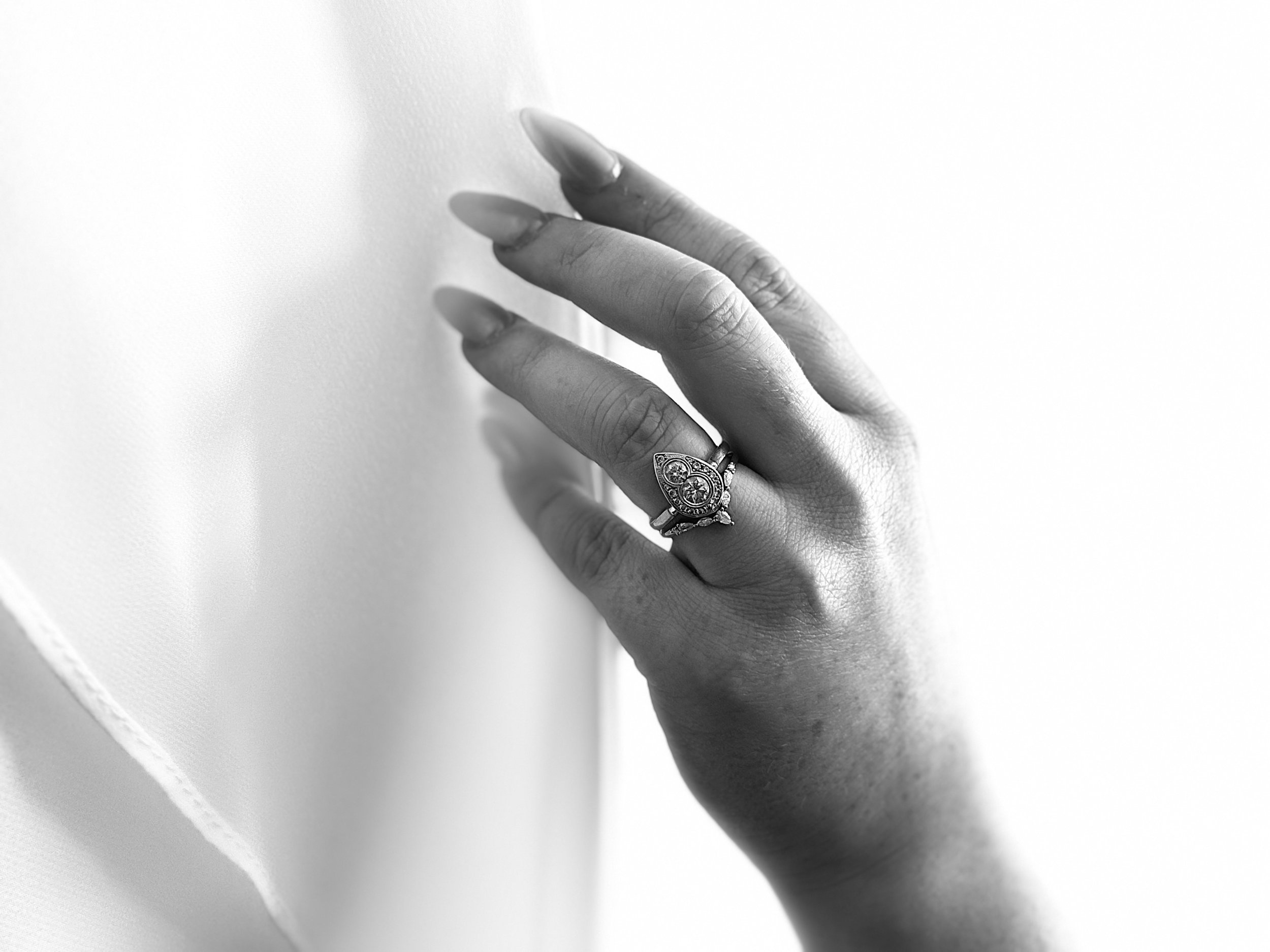 A close-up of a woman's hand with a diamond engagement ring on her finger, touching a plain wall. The image is black and white.