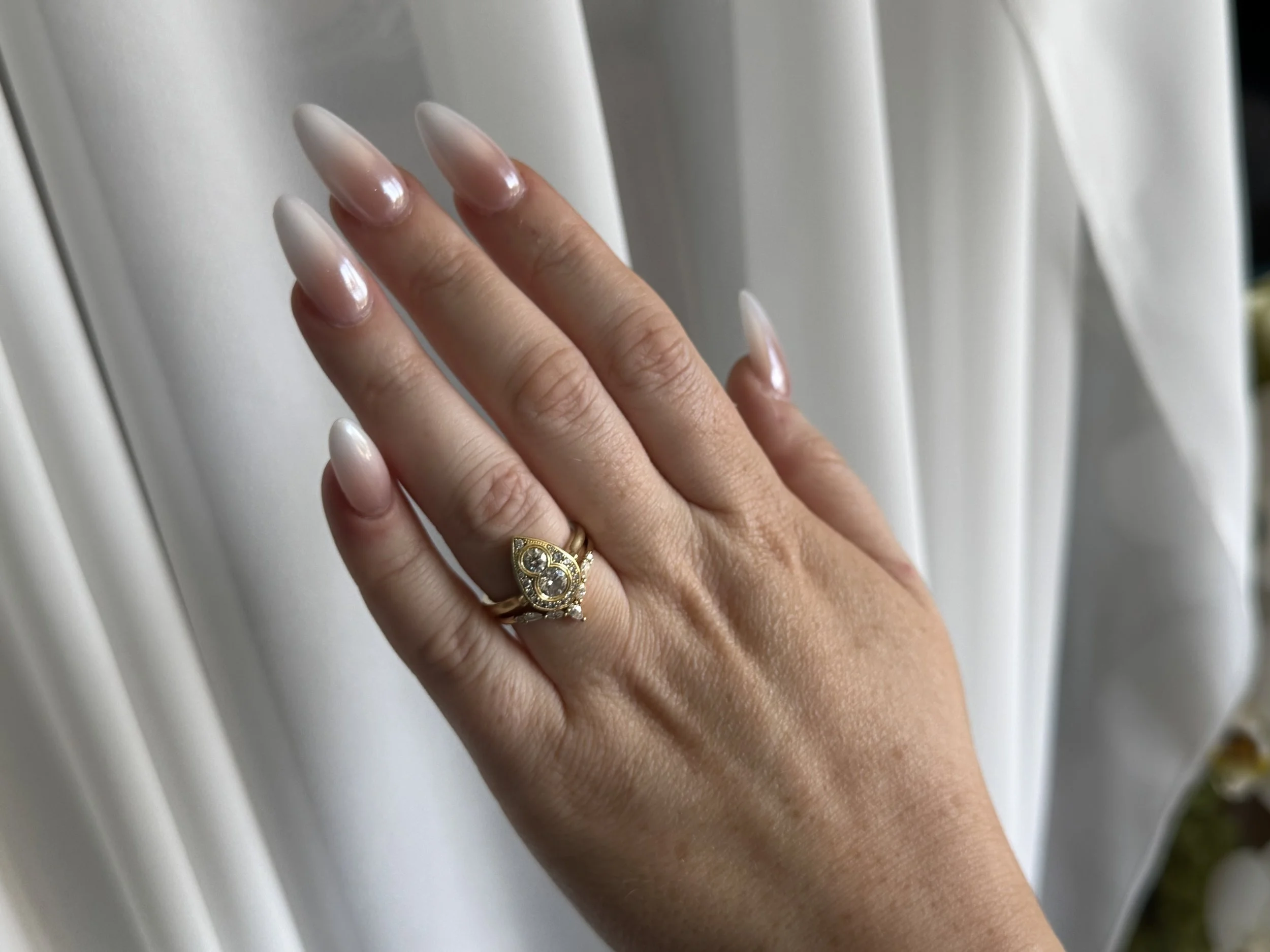 A woman's hand with long, manicured nails and a gold ring with diamonds, resting on a white surface.