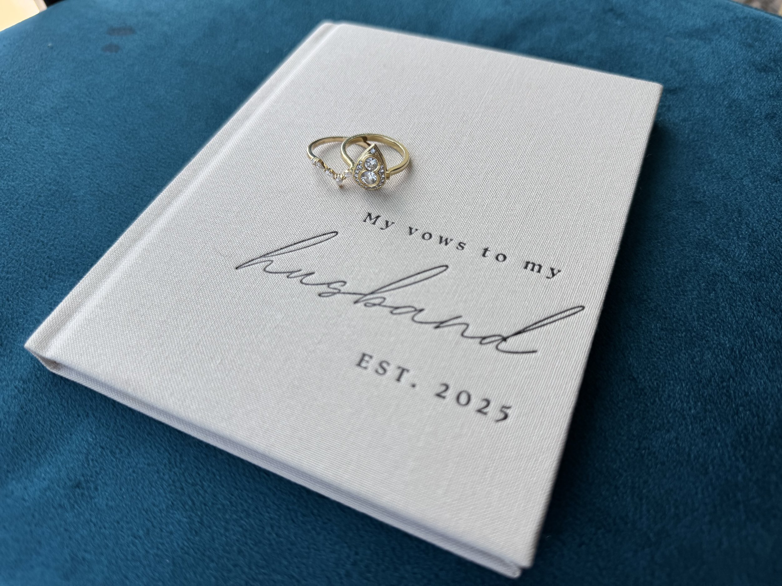 A white notebook with the phrase 'My vows to my fiancé' and 'EST. 2025' written on it, placed on a teal fabric surface. Two jewelry rings rest atop the notebook.