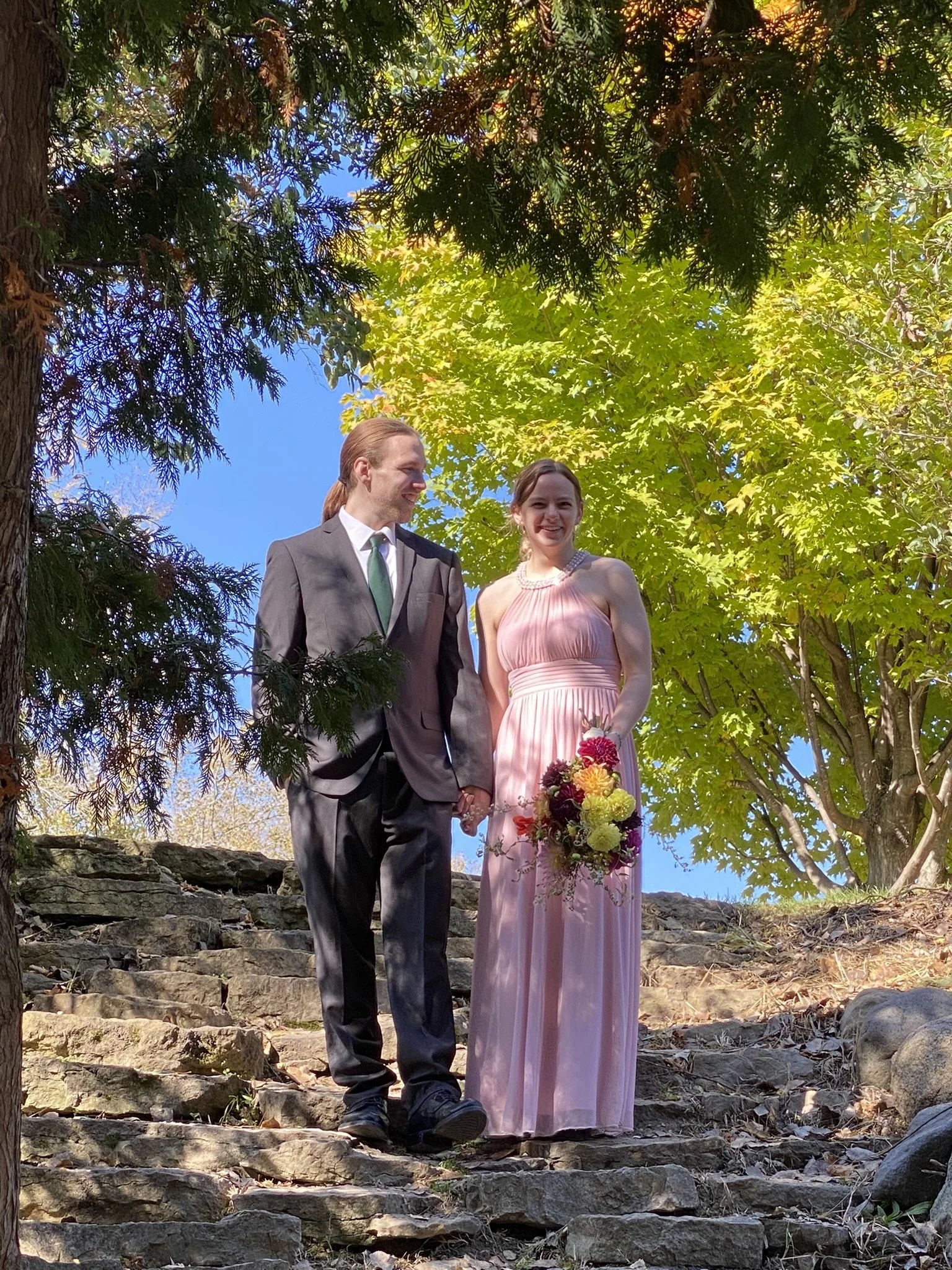 A couple standing outdoors on stone steps surrounded by trees, holding hands, dressed in formal attire for a wedding or special event, with the woman holding a colorful bouquet of flowers.