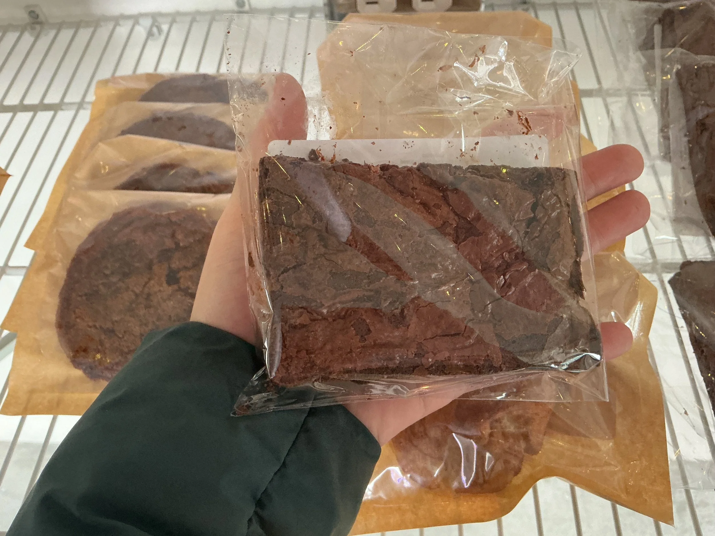 A person holding a sealed package of sliced beef jerky in a store. Other packages of beef jerky are visible on a shelf beneath a paper or cardboard backing.