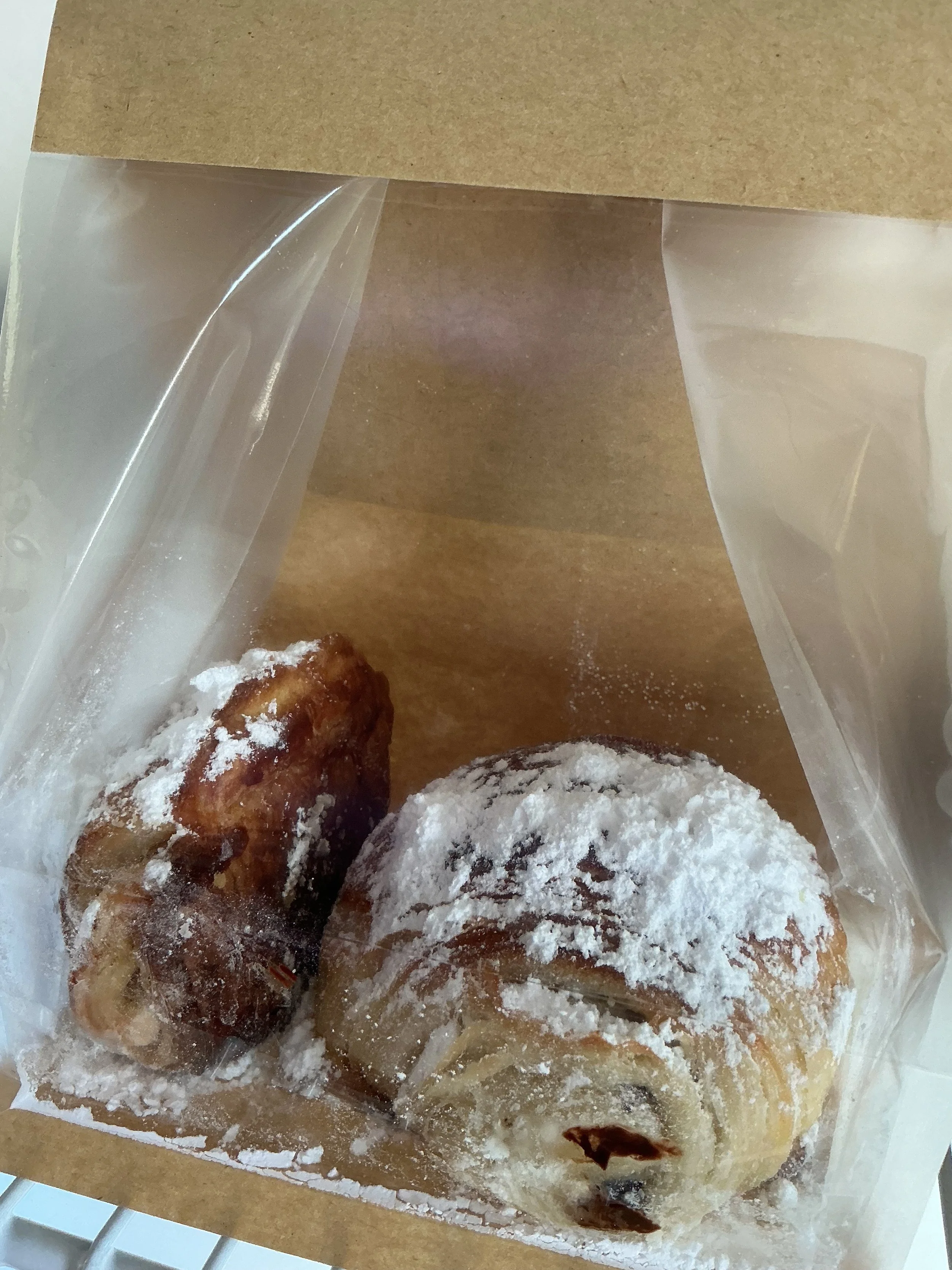 Two powdered sugar-covered baked goods inside a clear plastic bag, with a brown paper top, on a white surface.