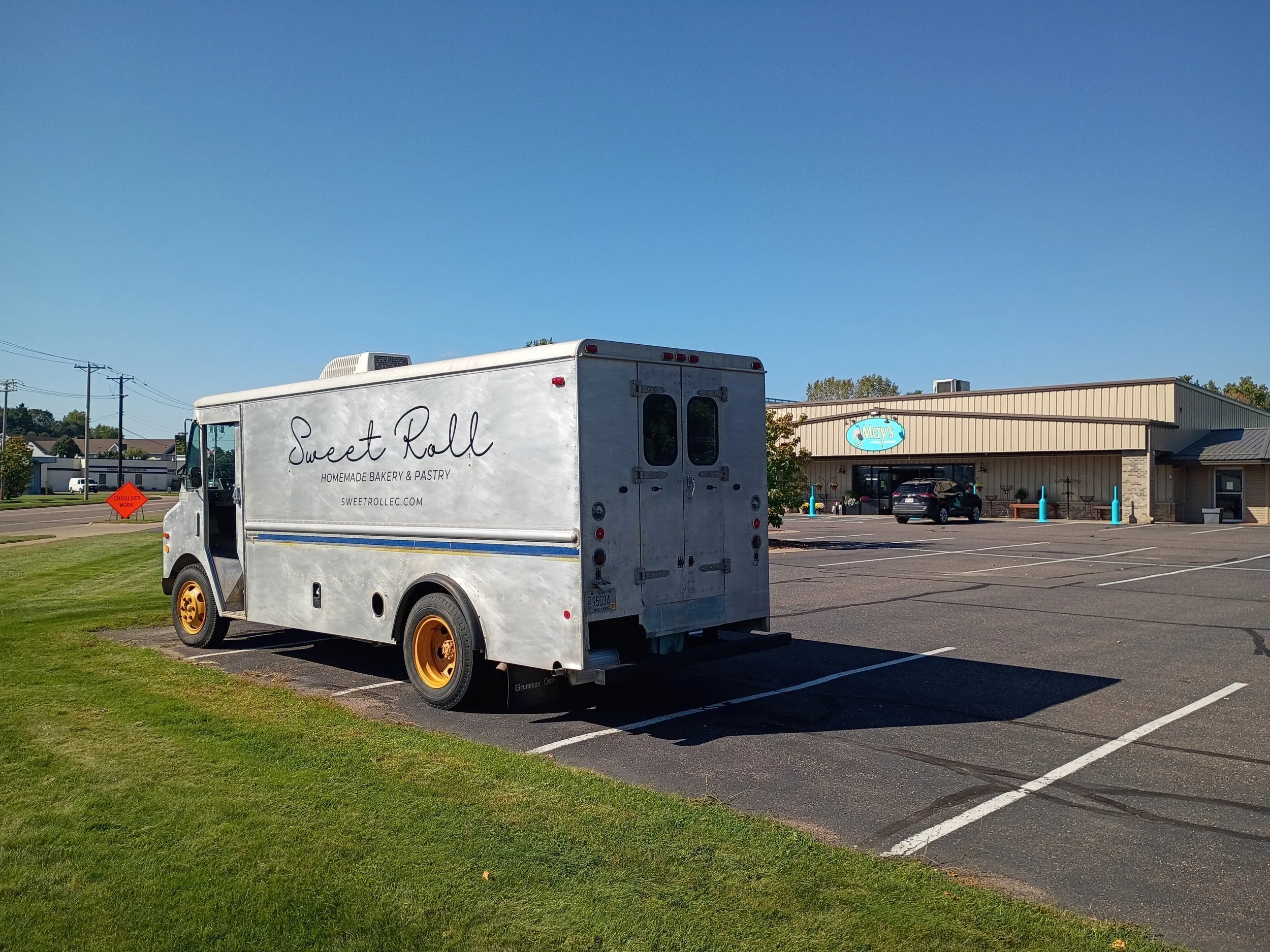 A food truck labeled 'Sweet Roll Homemade Bakery & Pastry' is parked in an empty parking lot near a building with a sign that reads 'Mays' on a clear day with blue skies.