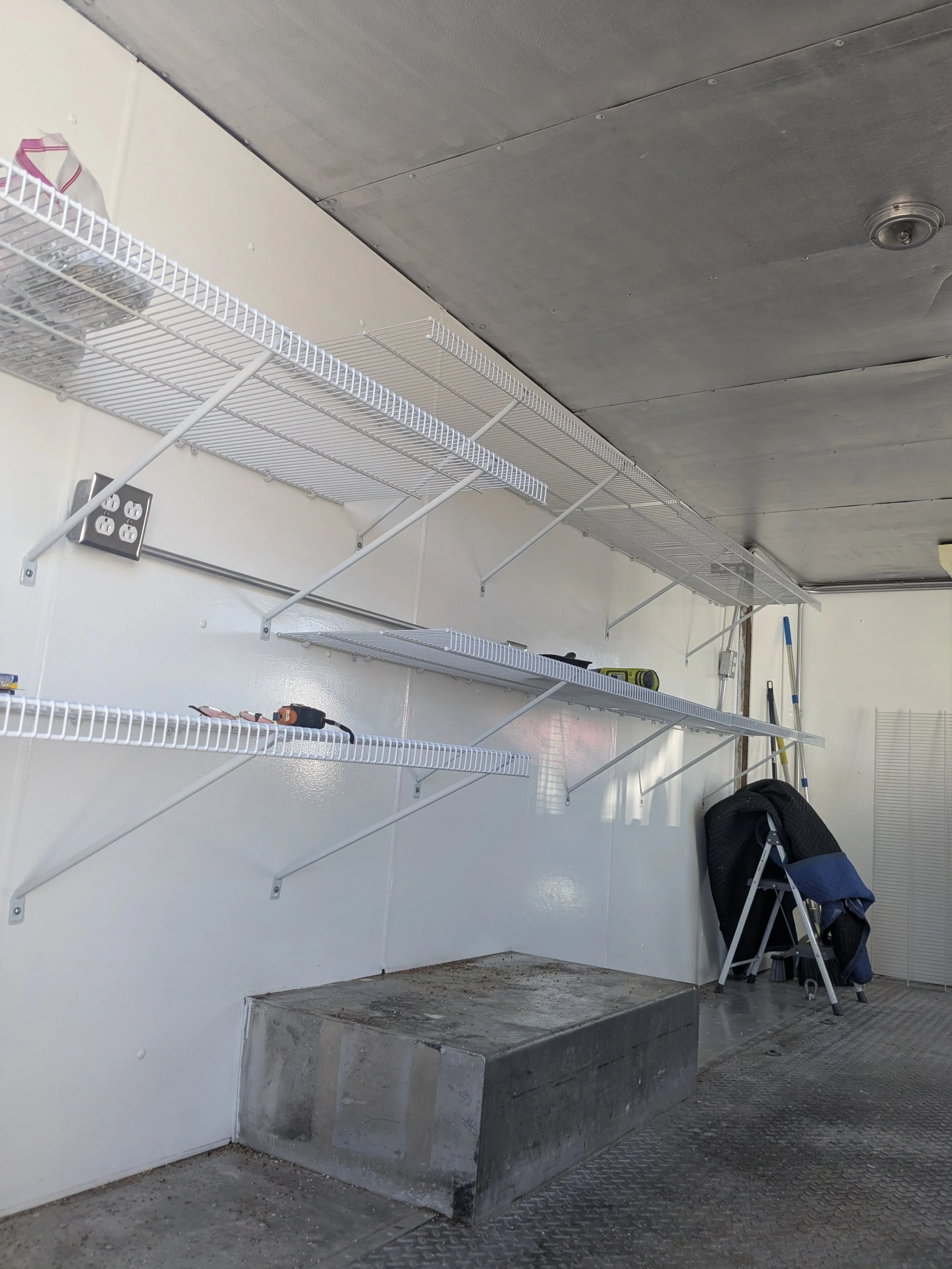 Empty white wire shelves mounted on a wall in a garage or storage room. A folded chair, some tools, and a broom are leaning against the wall. The floor is concrete with some dirt, and overhead lighting is visible.