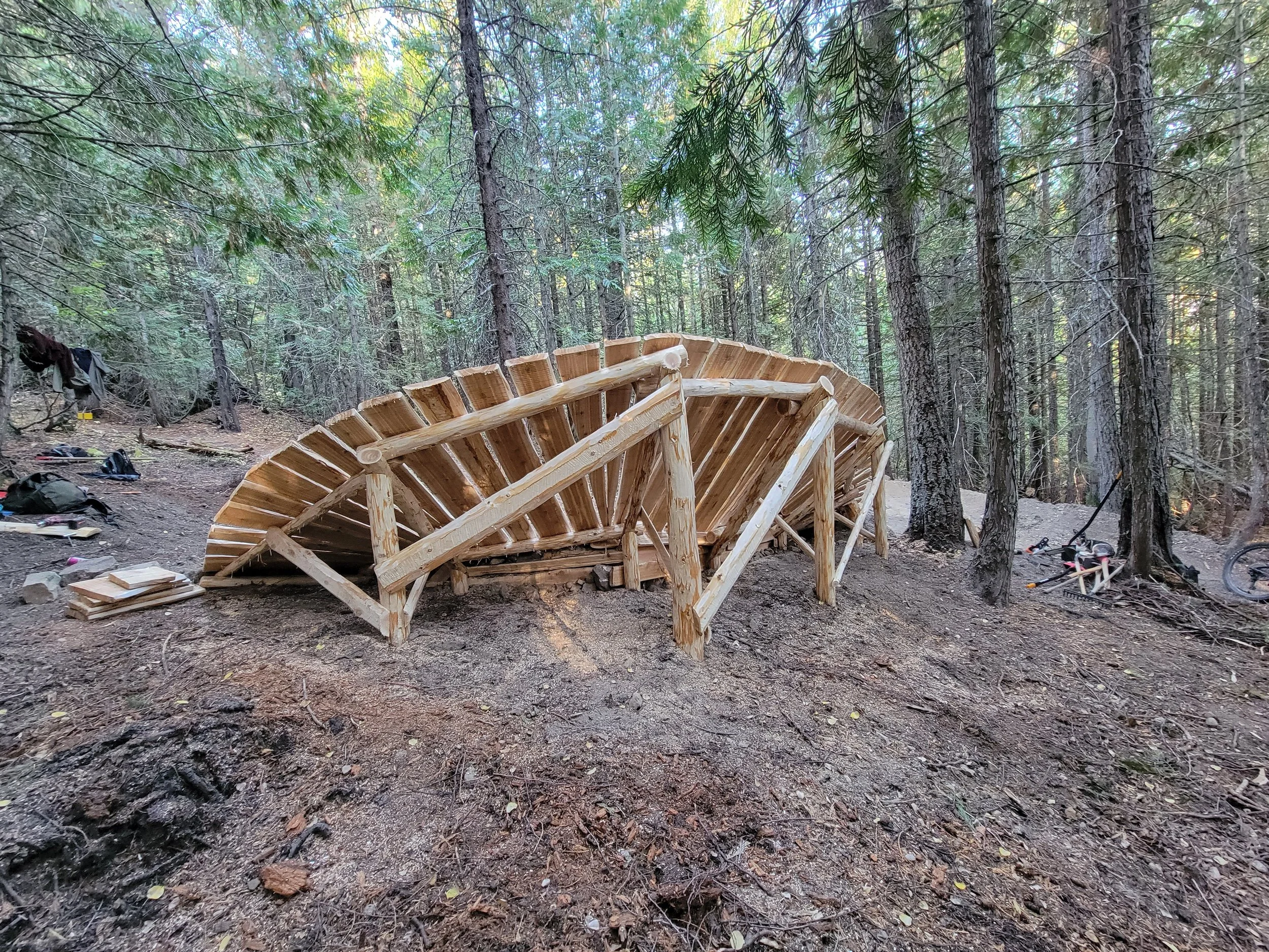 A partially built wooden structure in a forest, surrounded by trees and construction tools on the ground.