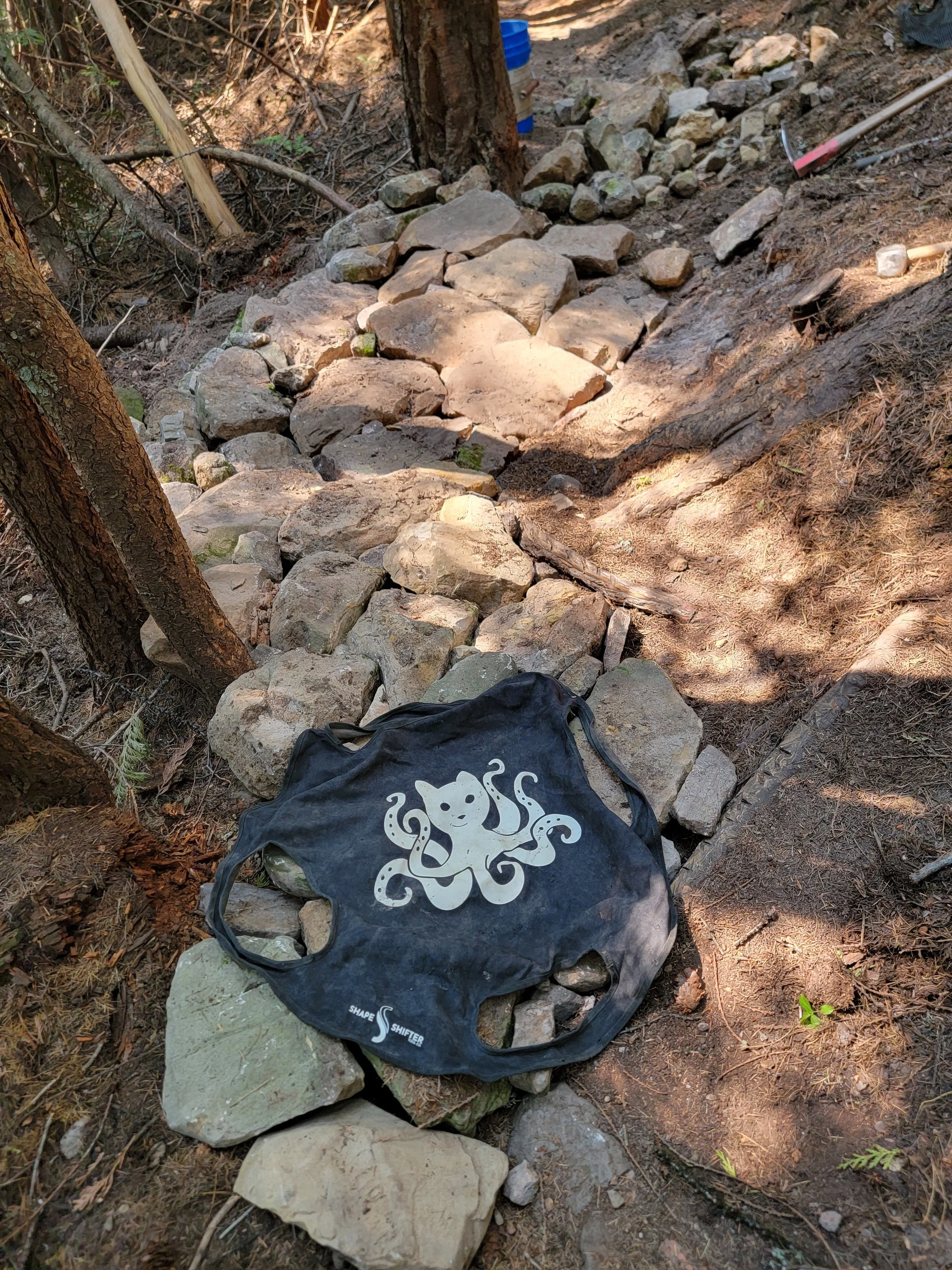 A rocky trail under trees with a black duffel bag featuring an octopus illustration resting on the rocks.