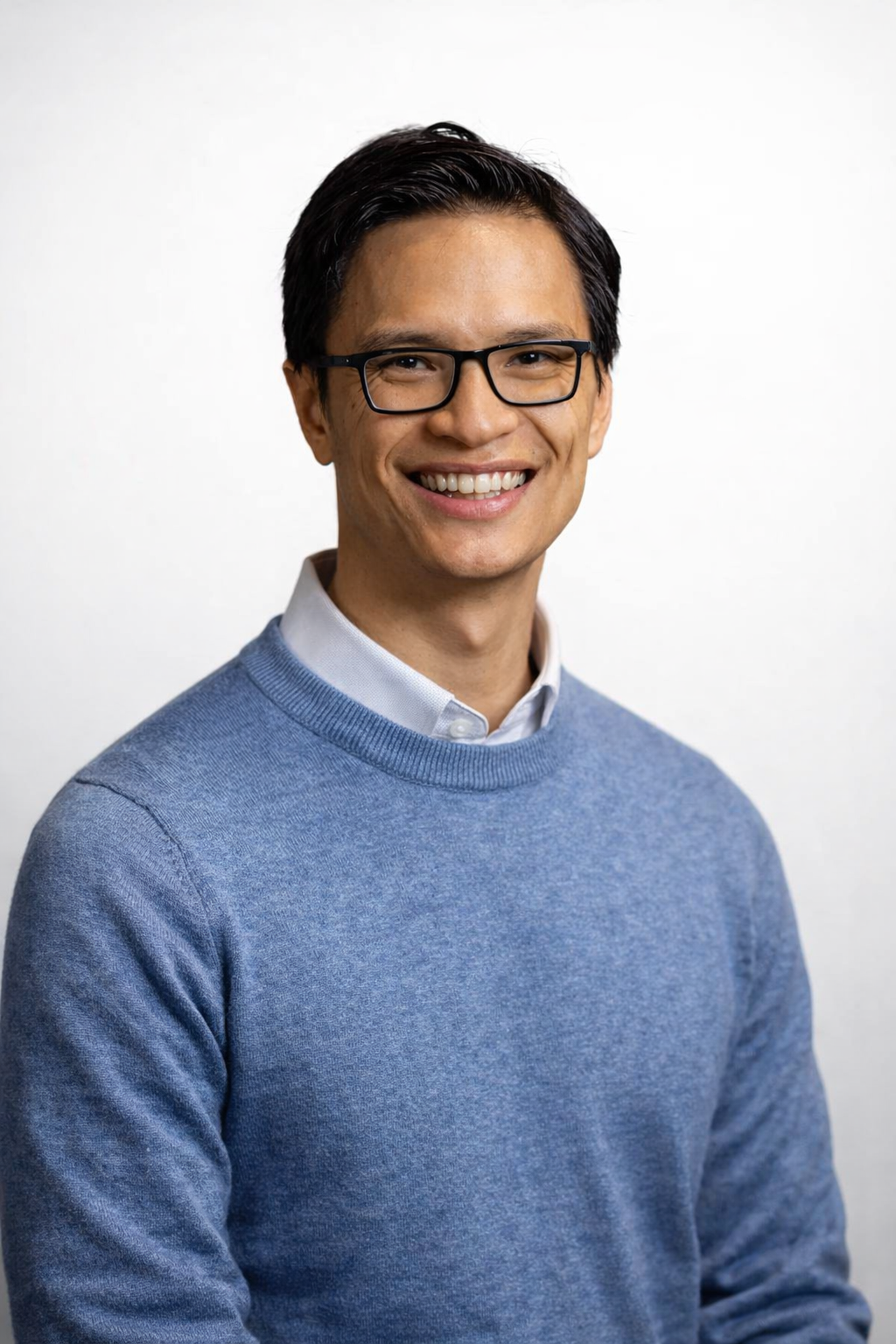 A smiling man with dark hair, glasses, wearing a light blue sweater over a white collared shirt, standing against a plain white background.