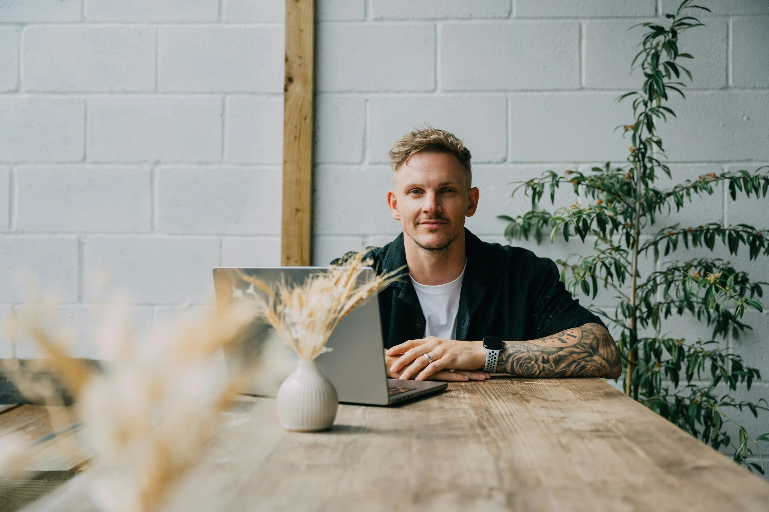 A man with tattoos and a smartwatch sitting at a wooden table with a laptop, in front of a white brick wall and a tall green plant.