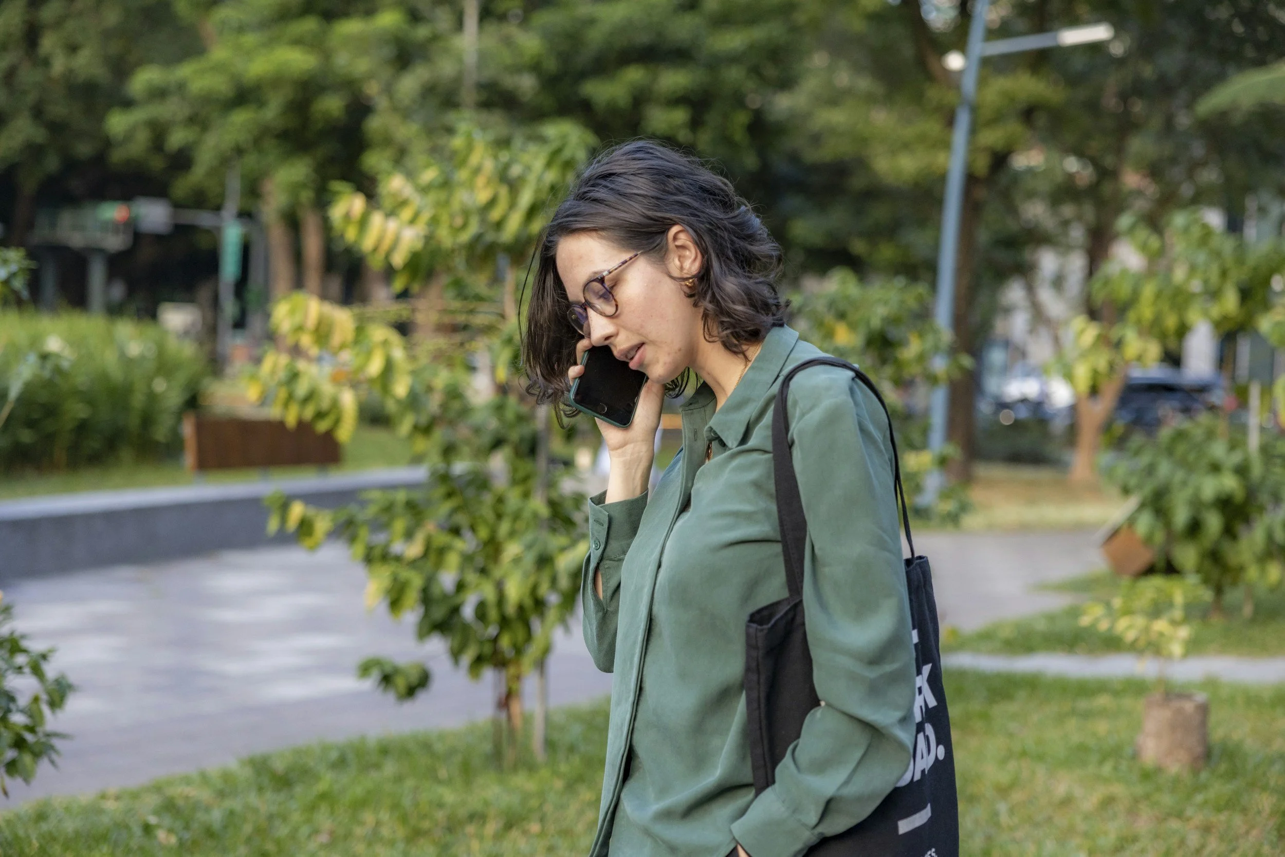 A woman with glasses and dark wavy hair speaking on her phone outdoors in a park with green trees and grass.