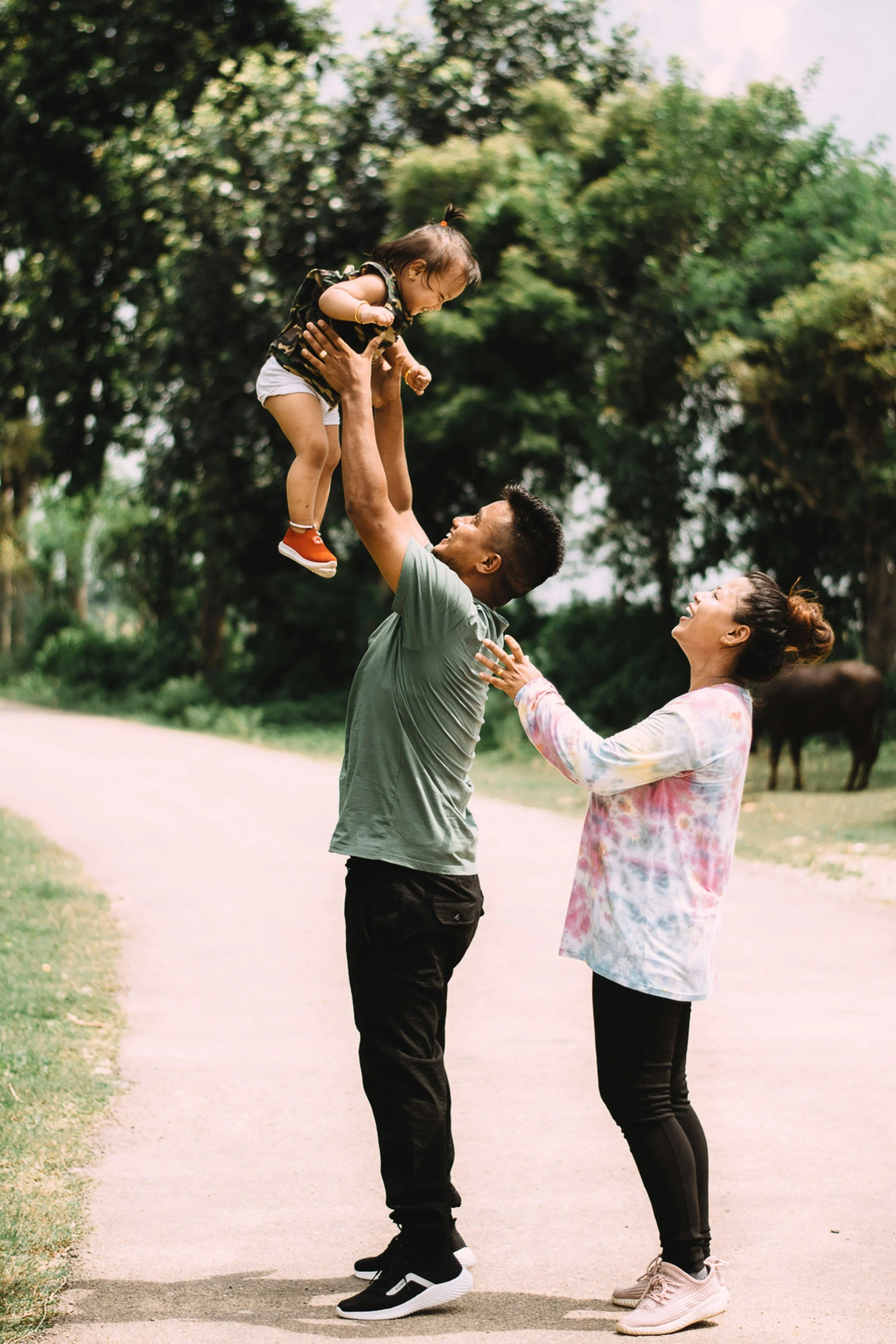 A family enjoying time outdoors on a paved trail surrounded by trees. A man is lifting a young girl into the air while a woman watches and smiles nearby.