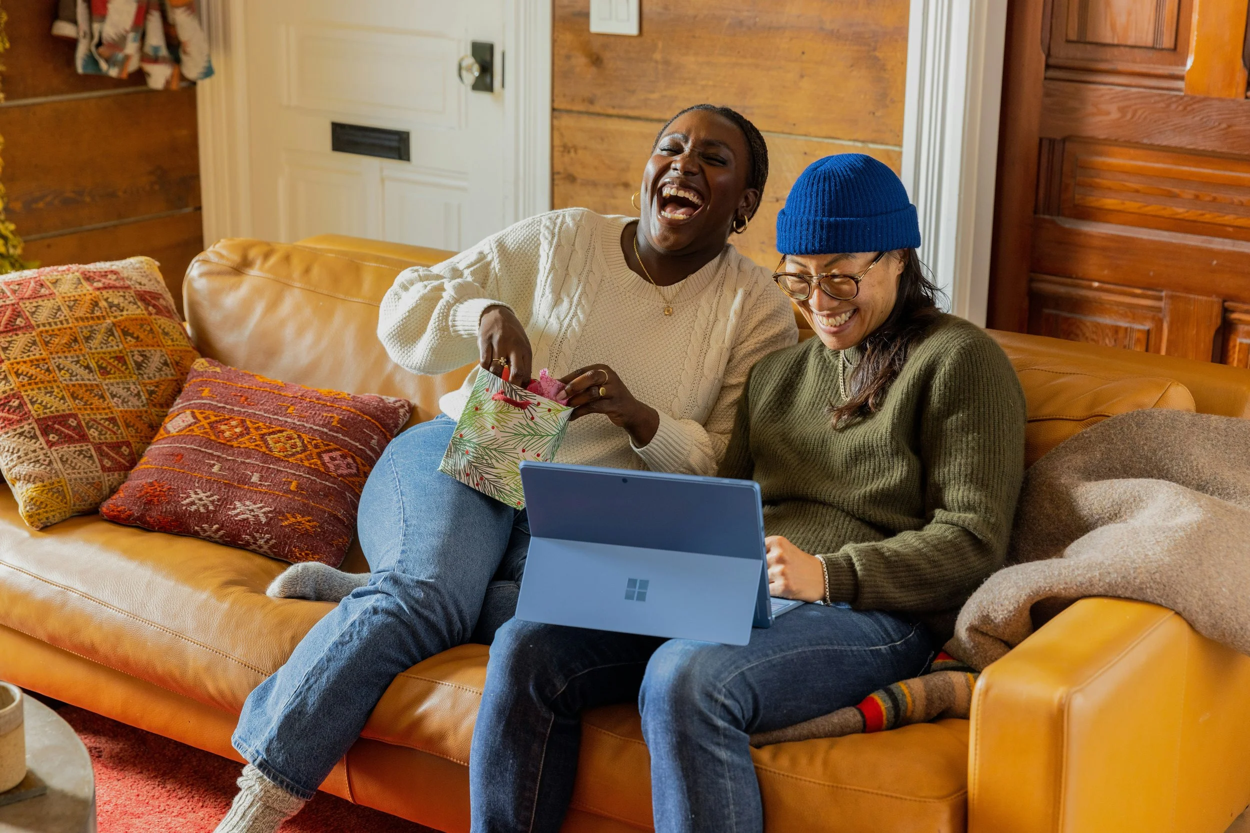 Two women sitting on a tan leather couch, exchanging gifts and laughing, with a laptop in front of them in a cozy room with wood-paneled walls and colorful pillows.