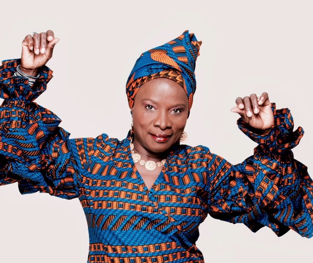 A woman wearing colorful traditional African attire, including a matching headwrap and dress, posing confidently with her fists raised.
