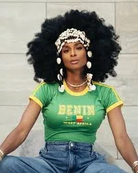 A woman with a large, curly afro hairstyle wearing a green t-shirt, sitting outdoors against a light-colored background. She has a white headscarf, large white earrings, and layered necklaces.